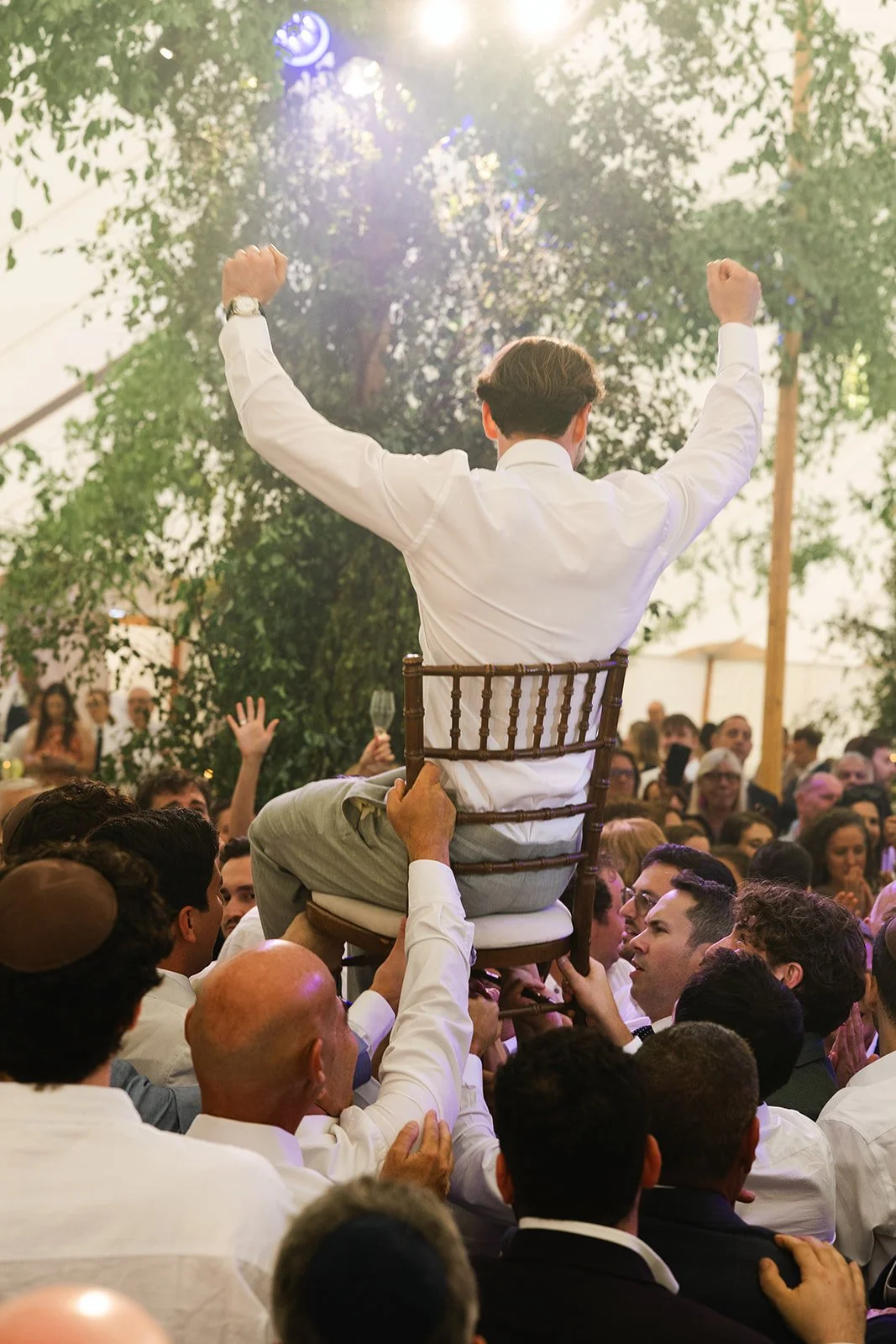 A man wearing a white shirt is seated on a chair held up by several people at a celebration or party. The man has his arms raised in a victorious gesture, and the crowd around him is lively with some people raising glasses, indicating a festive atmos