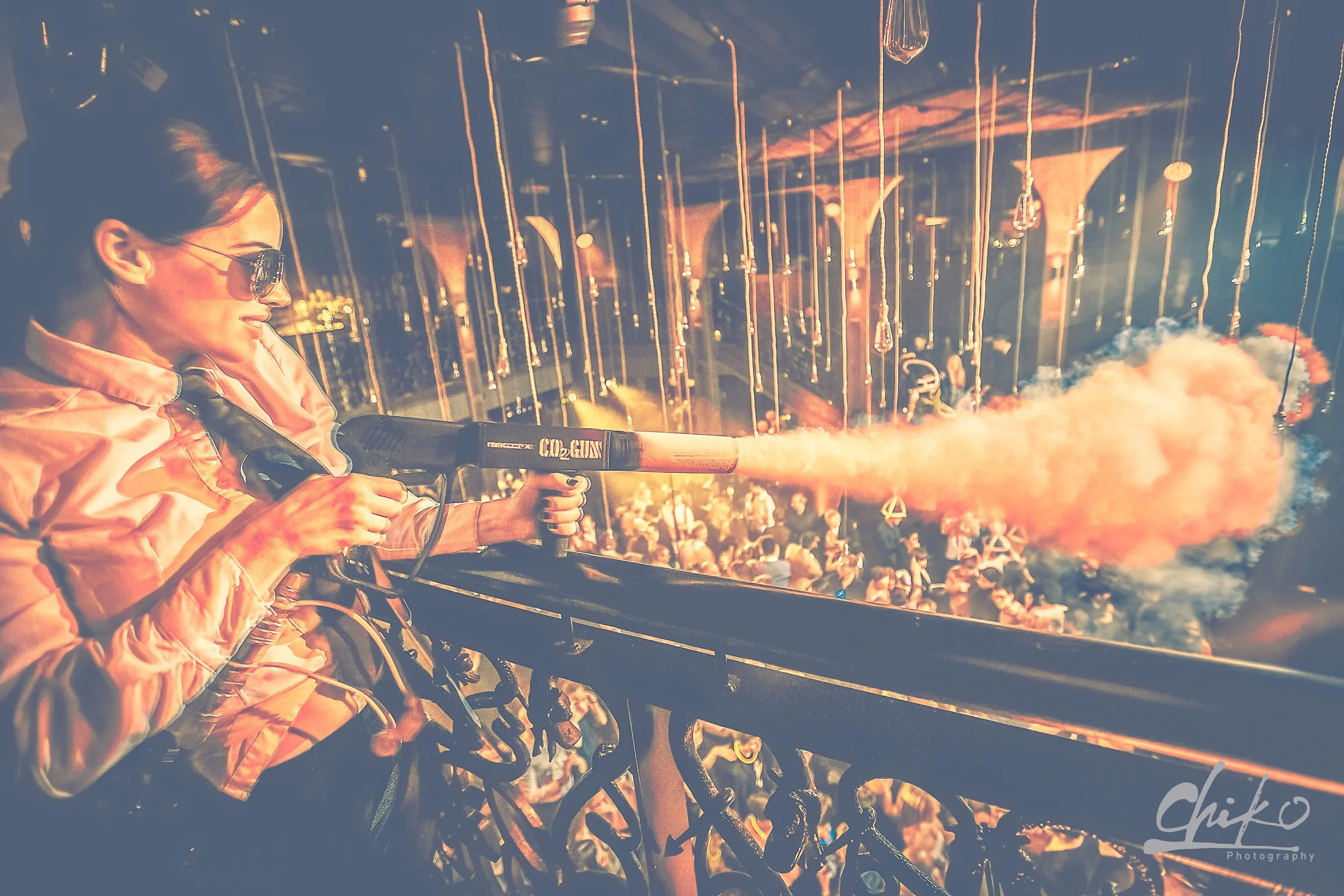 A woman wearing sunglasses and a peach-colored jacket using a handheld firework launcher during a firework display at night with many spectators and fireworks in the background.