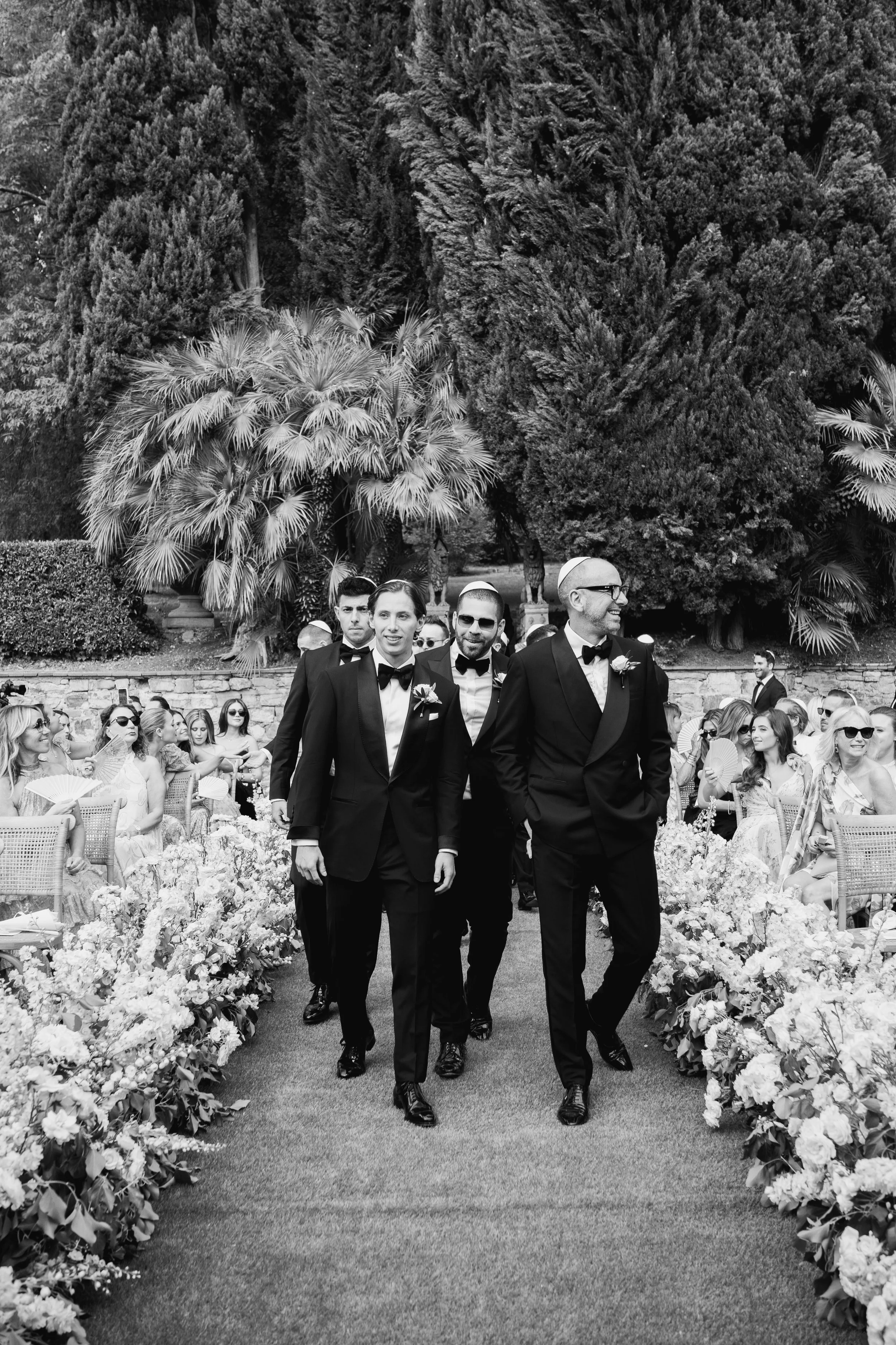 Men in tuxedos walking down an aisle at a wedding ceremony surrounded by floral arrangements and seated guests.