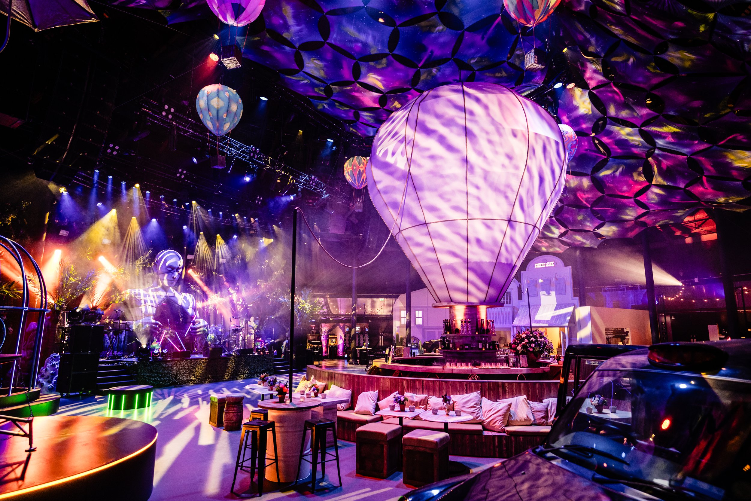 An indoor venue with colorful lighting, decorative hot air balloon sculptures, and a stage with musical performers. There are seating areas with sofas and tables, and vibrant, futuristic decor.