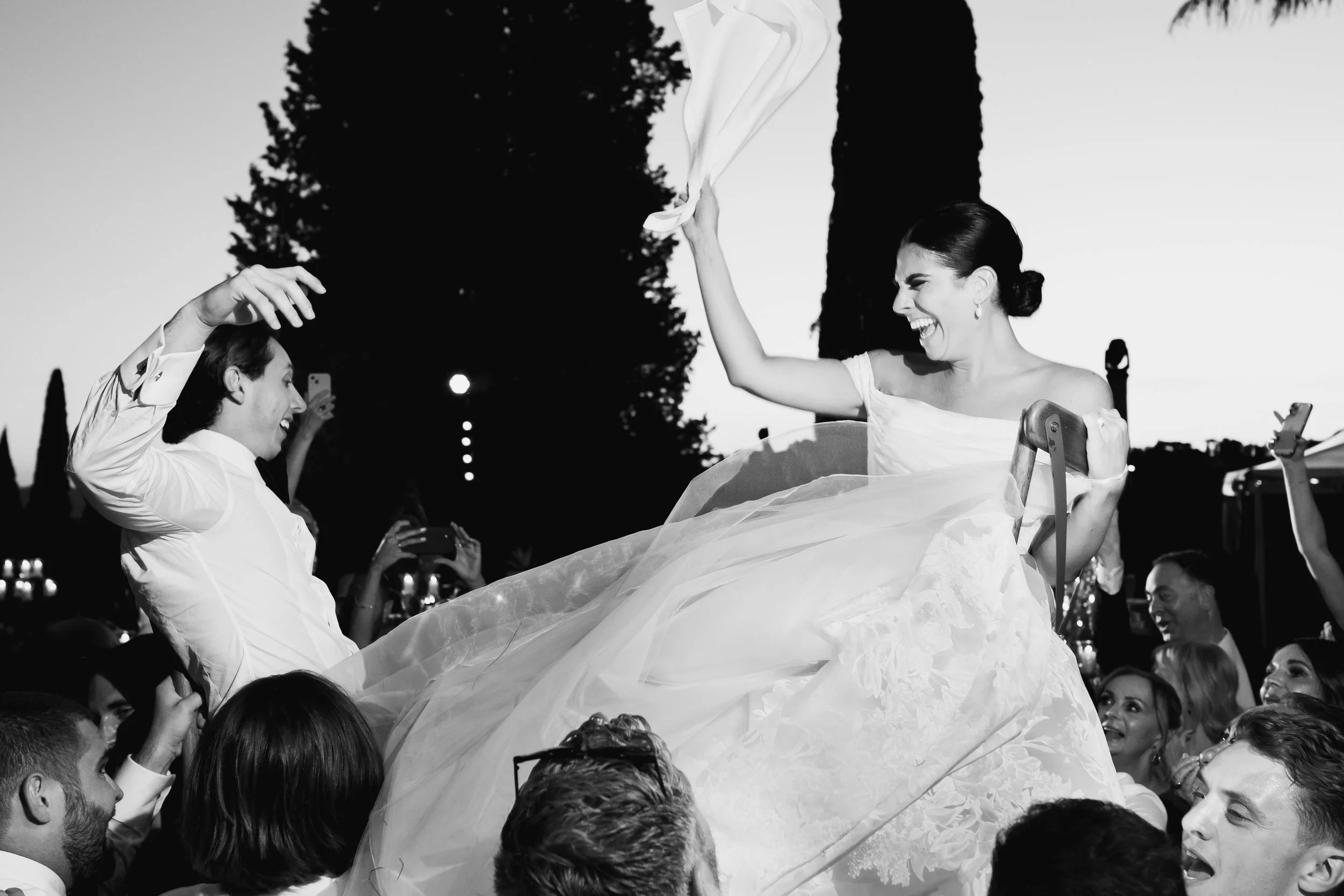 A woman in a wedding dress is being lifted above a crowd at an outdoor wedding smiling and holding a cloth in her hand, with people around taking pictures at Villa Palmieri in Florence