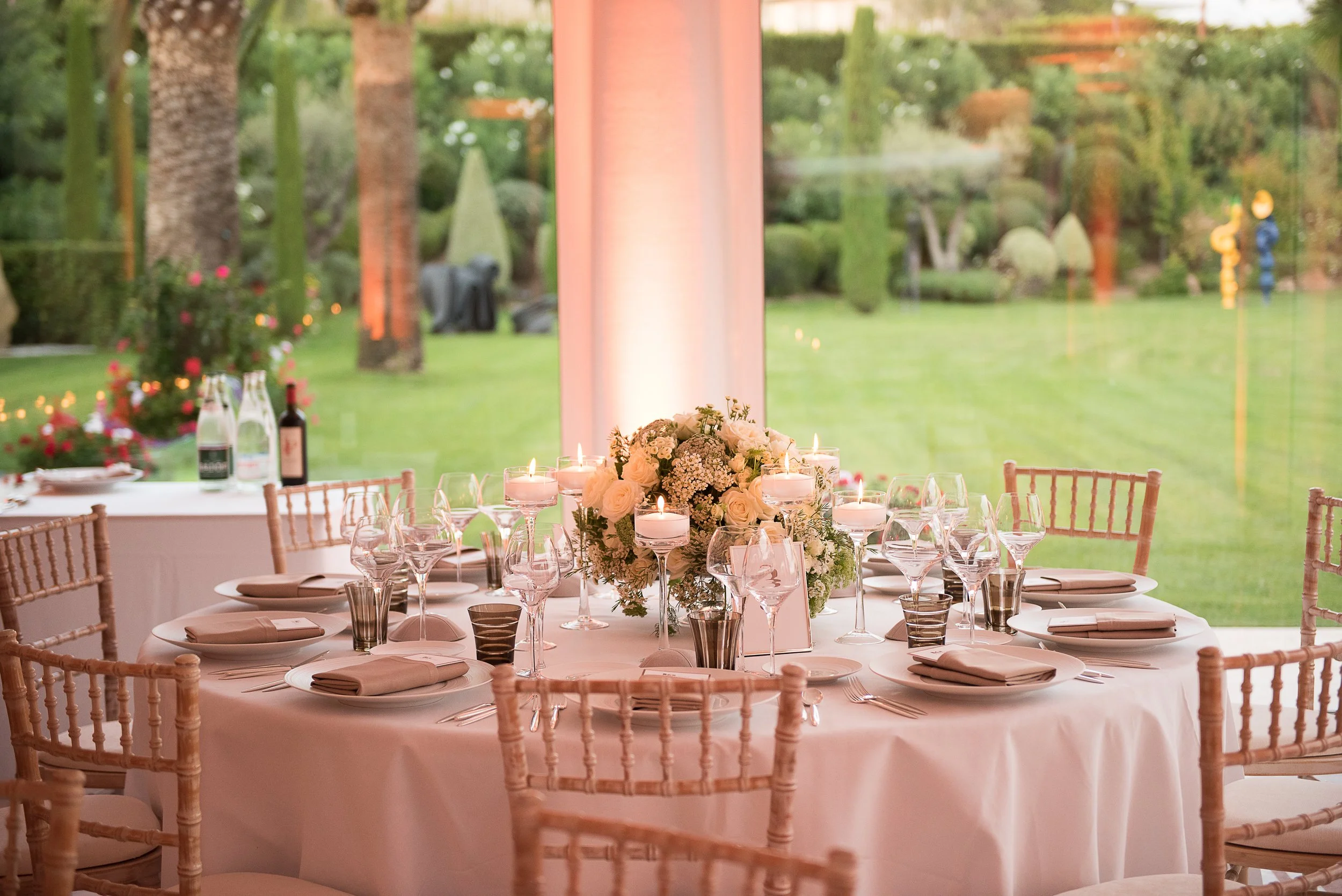 Elegant dining table set with white tablecloth, floral centerpiece with white roses, candles, wine glasses, and beige chairs, overlooking a lush green outdoor garden through large glass windows.