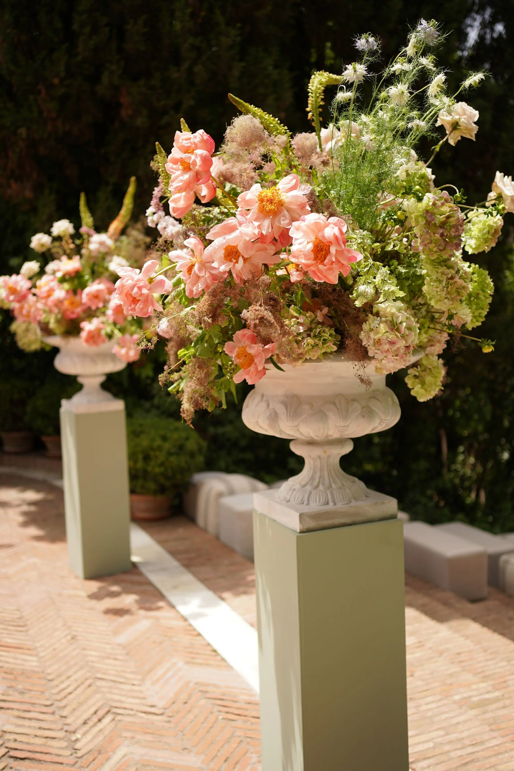 A floral arrangement with pink, white, and green flowers in a white urn on a pedestal outdoors, with a brick pathway and greenery in the background at Anantara Villa Padierna Resort in Marbella