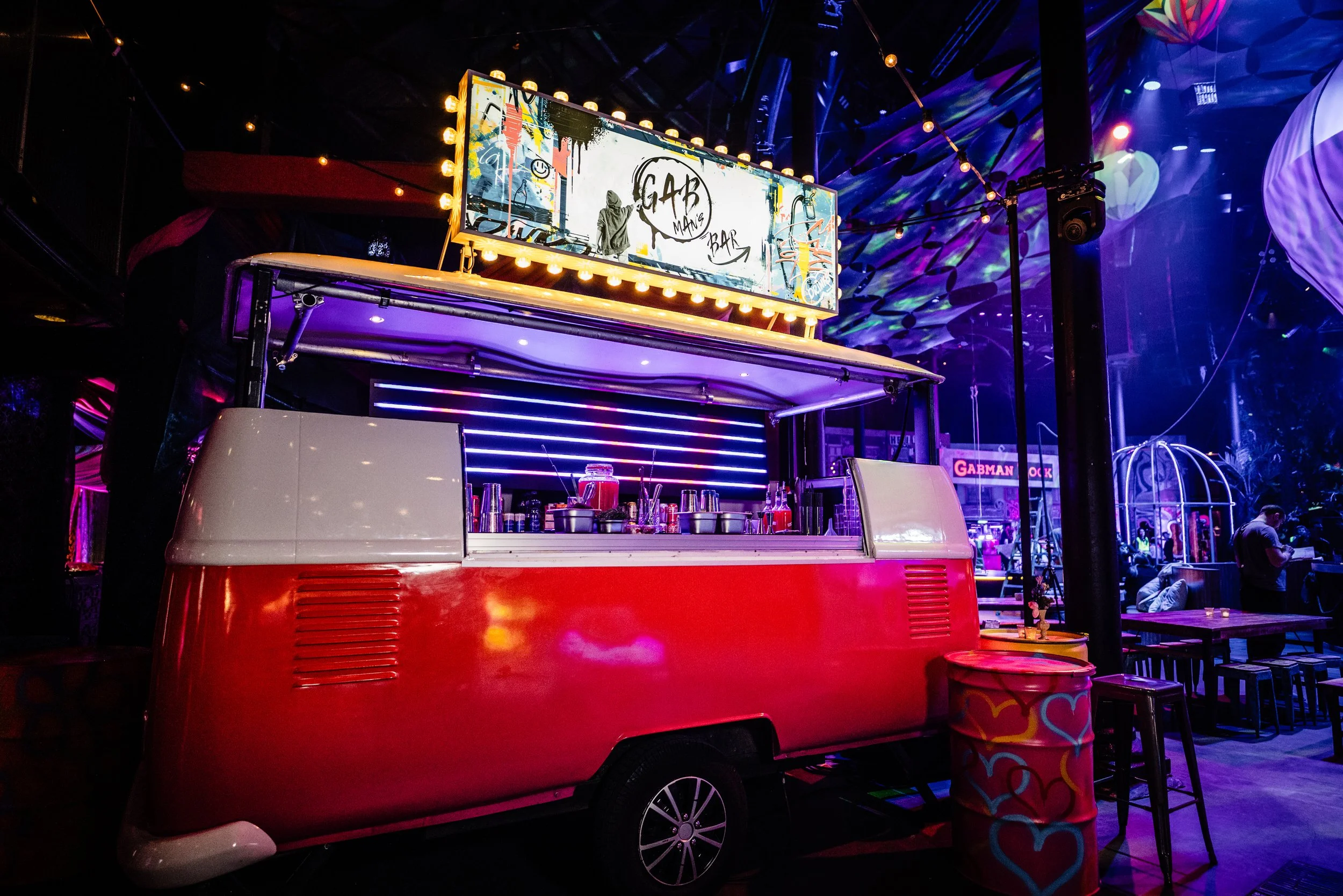 A red and white vintage ice cream truck set up as a food or drink stand inside a dimly lit, colorful venue with purple and blue lighting, decorative string lights, and a large electronic sign displaying graffiti-style art with the words 'GAB' and 'BA