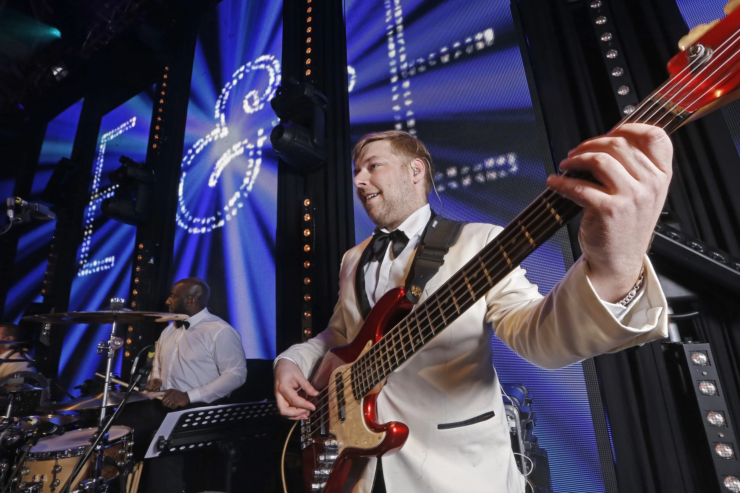A musician in a tuxedo jacket playing an electric guitar on stage with bright blue LED screens in the background and stage lights overhead.