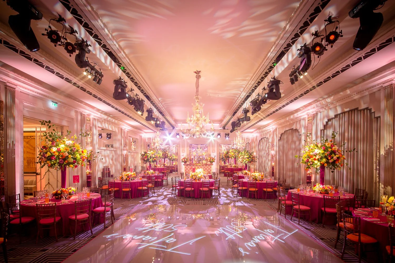 Elegant banquet hall decorated for a wedding reception with pink-themed table settings, large floral arrangements, chandeliers, and stage lighting.