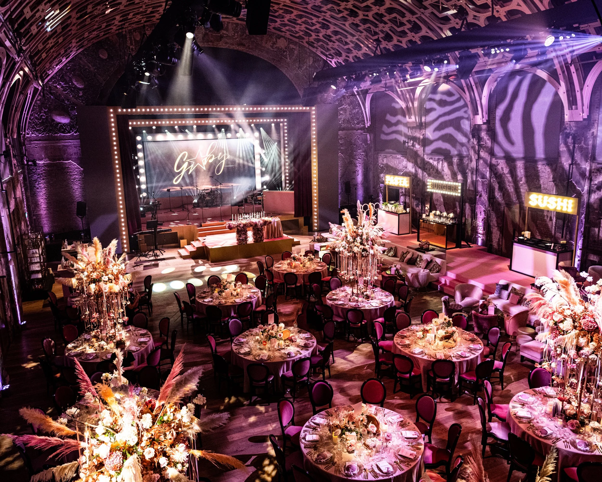 Elegant event space decorated with pink and purple lighting, round tables with floral centerpieces, a stage with musical instruments, and neon signs displaying food options like burgers, sushi, and pizza.