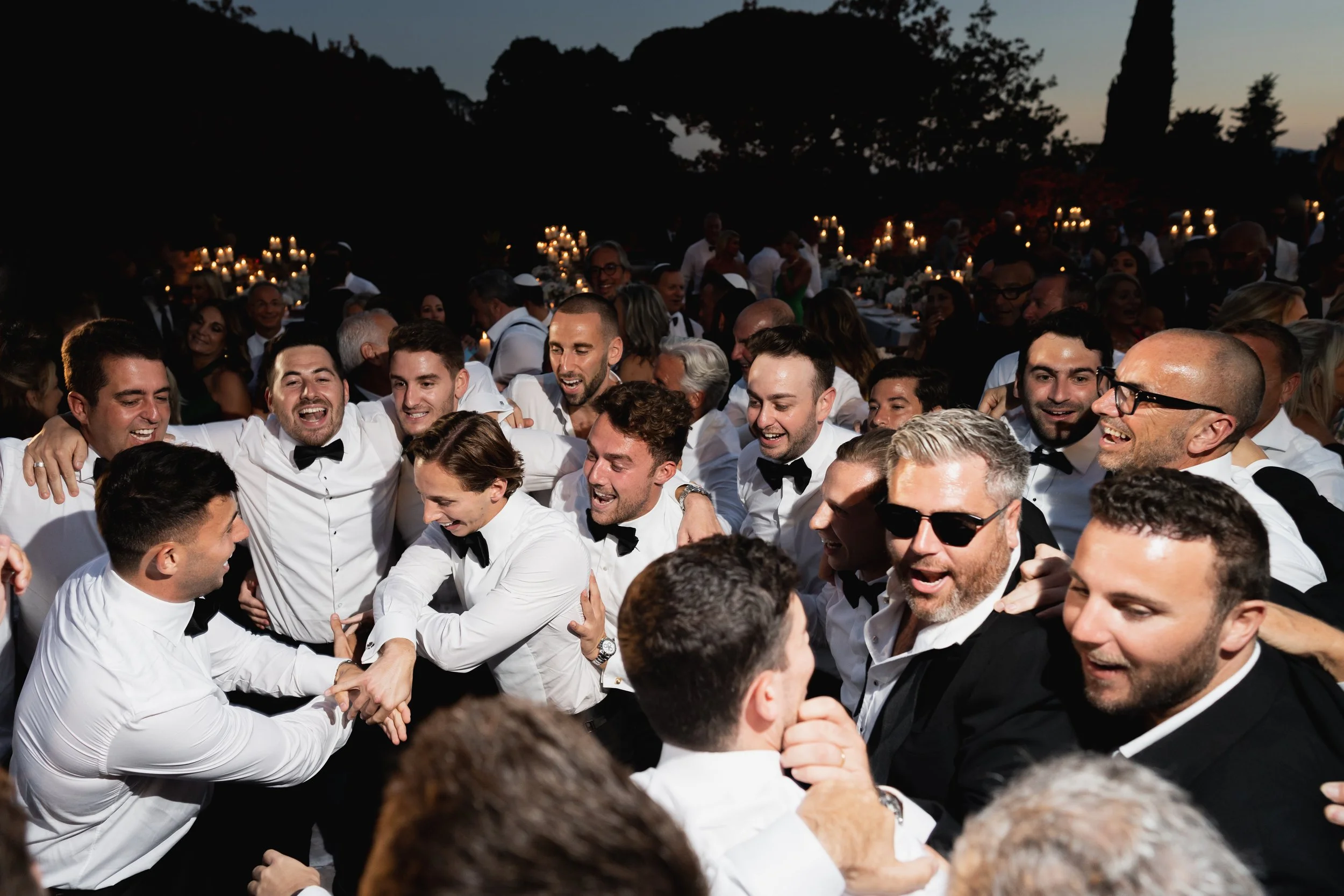 A large group of people dressed in formal attire celebrating outdoors with many smiling and engaging in lively conversation and handshakes. Jewish tradition