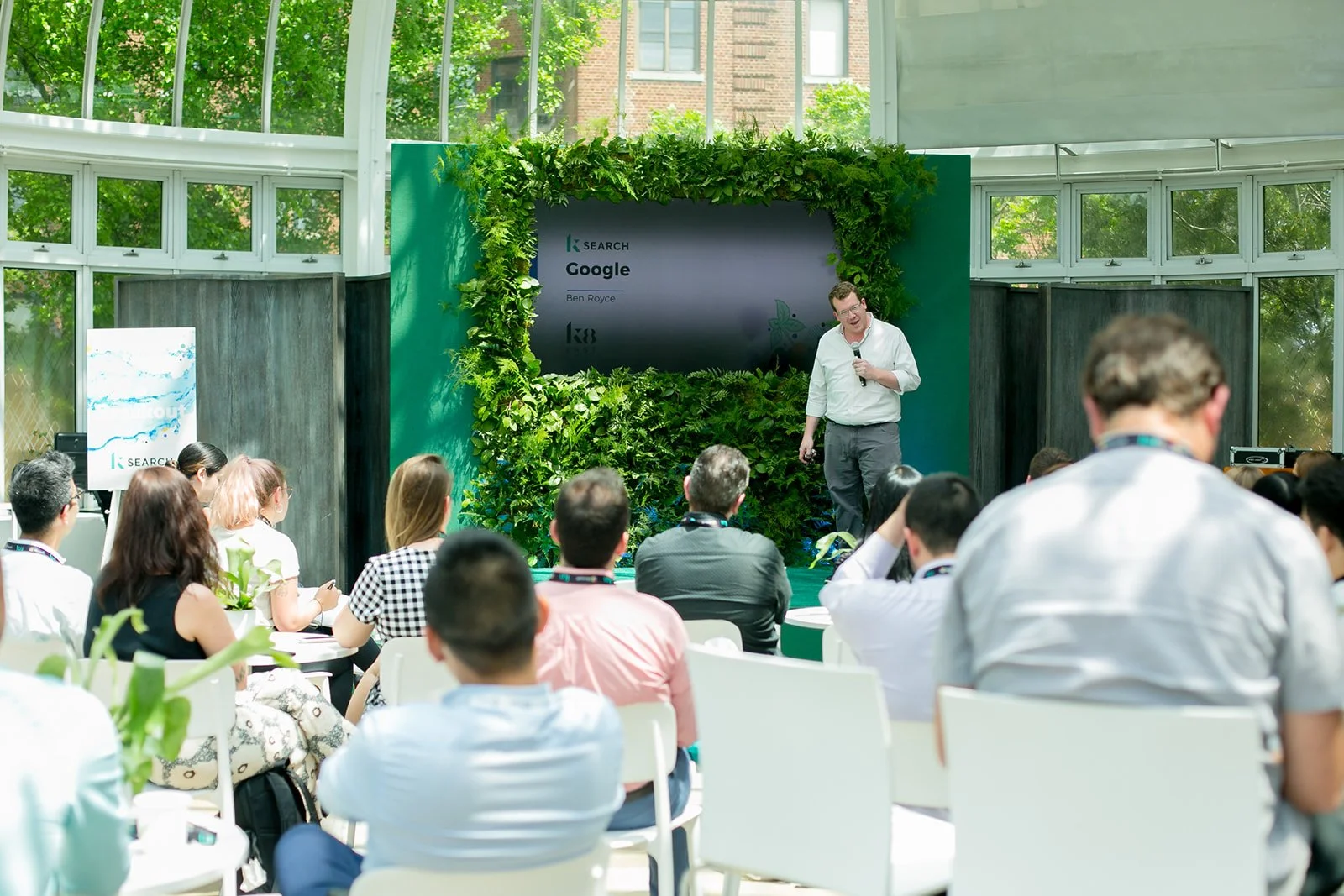 A man in a white shirt giving a presentation to an audience in a bright room with large windows and green plants, with a screen displaying the Google search logo and the presenter’s name.