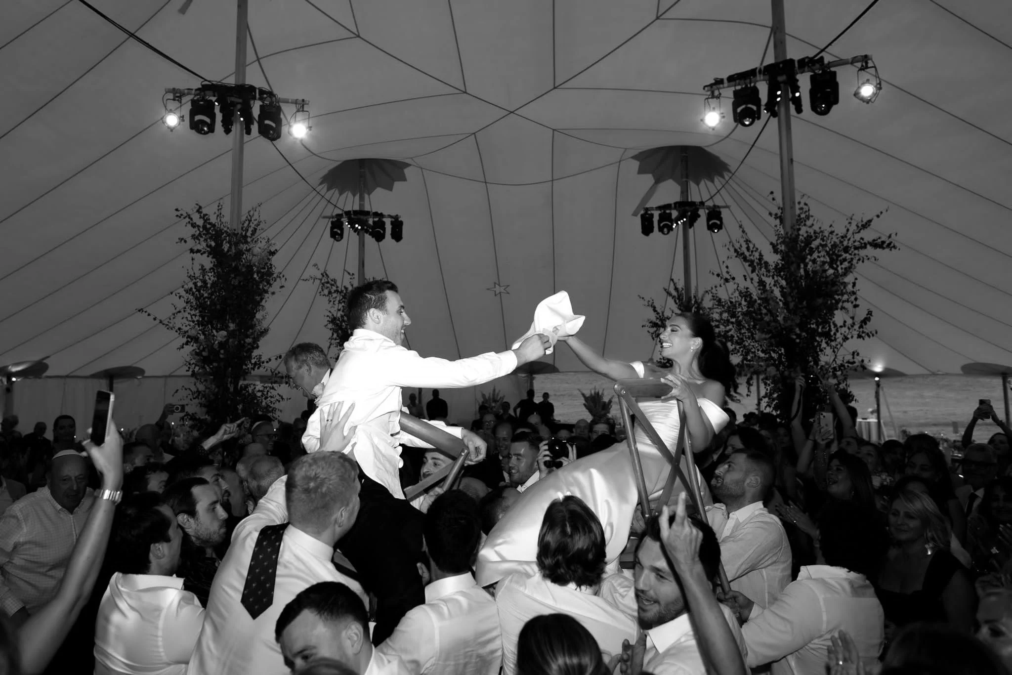 A couple dancing at a wedding reception in a large decorated tent filled with guests. The groom and bride are smiling and holding hands. The scene is lively with many people and some are taking photos.