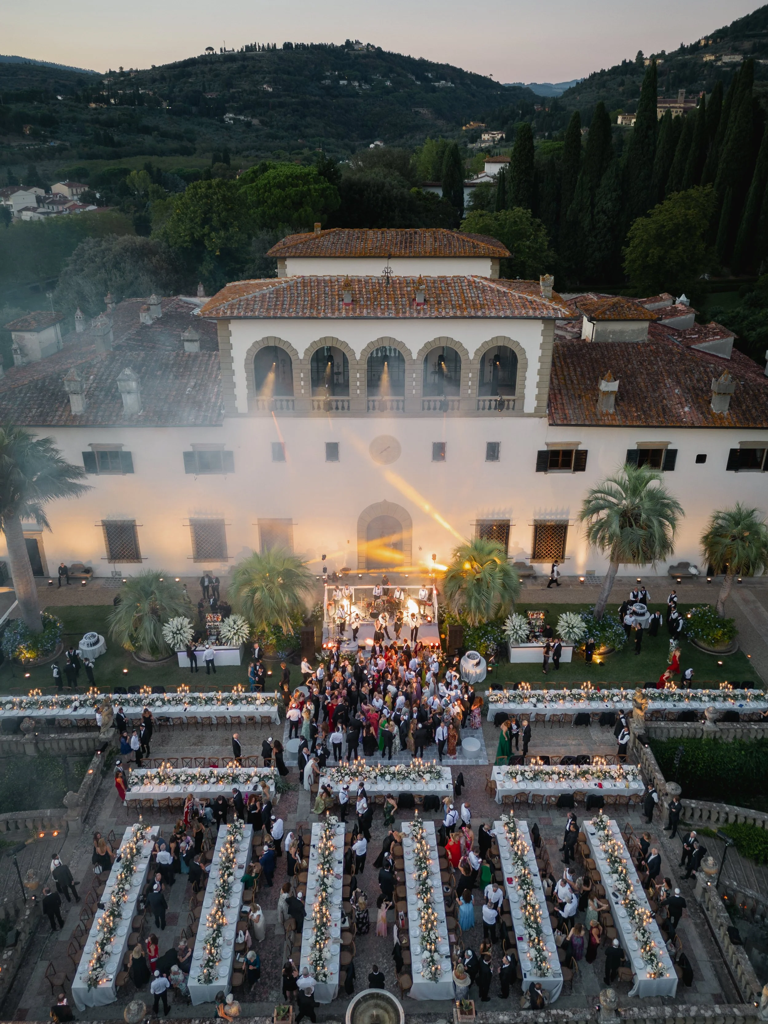 Florence Outdoor wedding reception at Villa Palmieri at sunset with long banquet tables decorated with candles and flowers, surrounded by guests, palm trees, and a historic building with a tiled roof in a hilly landscape.