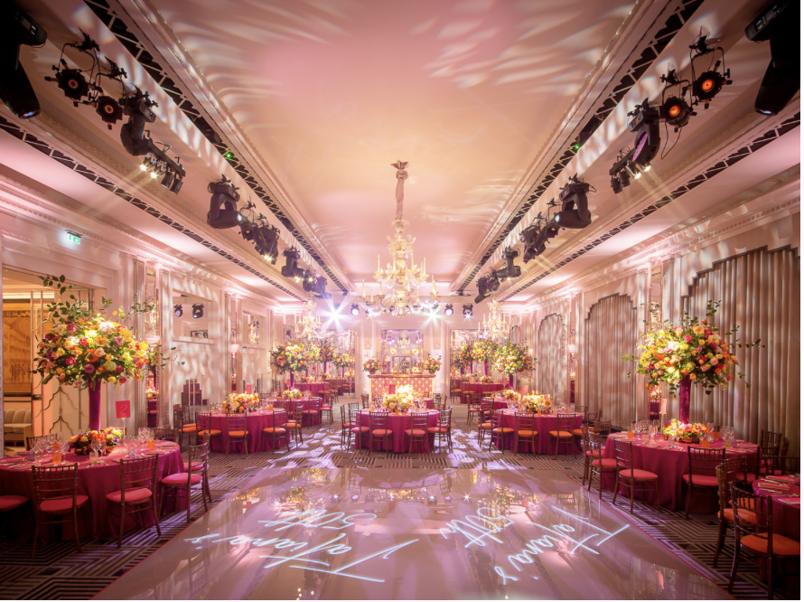 Elegant banquet hall decorated with pink tablecloths, large floral centerpieces, and a crystal chandelier, with dance floor and lighting effects.