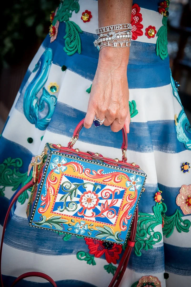 A woman holding a colorful, decorative handbag. She is wearing multiple silver bracelets and a large ring. The dress features blue and white horizontal stripes with floral embroidery and embellishments.