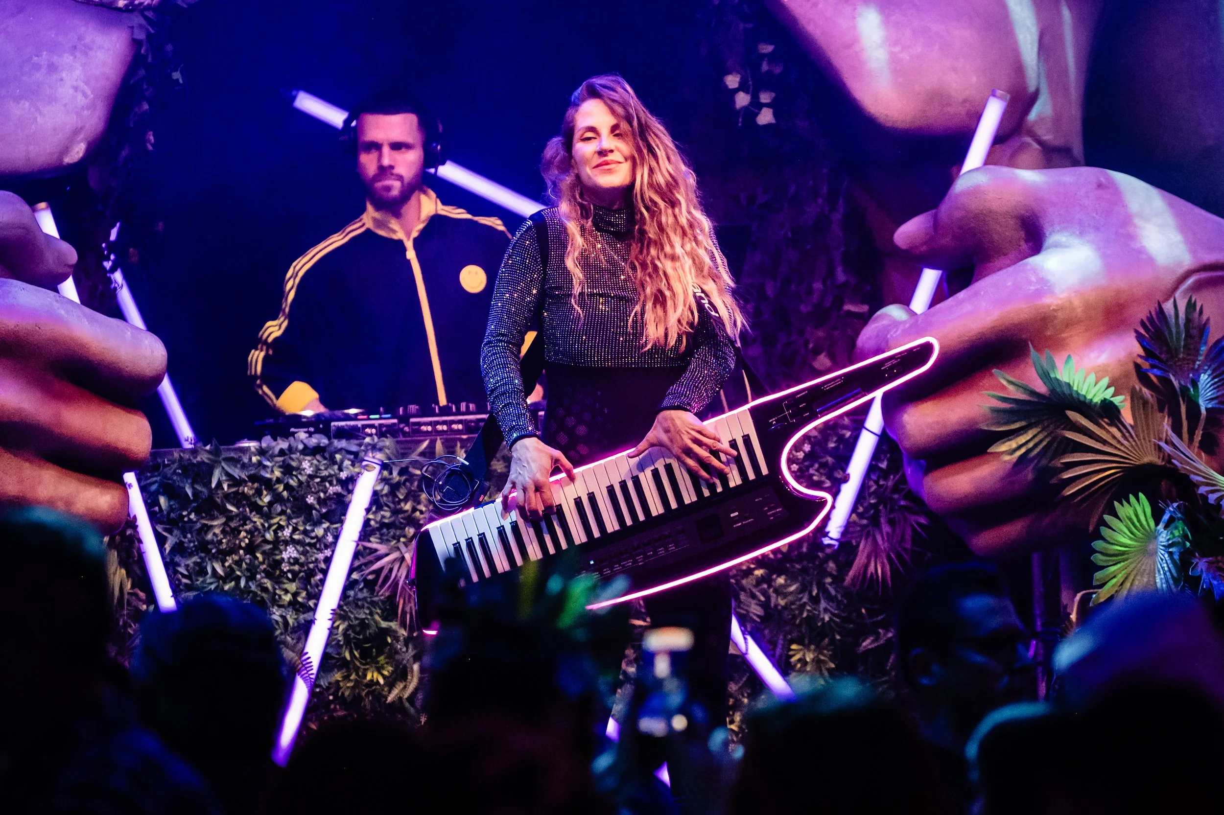 A woman with long curly hair playing a keyboard with neon purple lights, accompanied by a man with headphones and a serious expression in a dark, colorful, and vibrant setting with large plant-like structures and glowing elements.