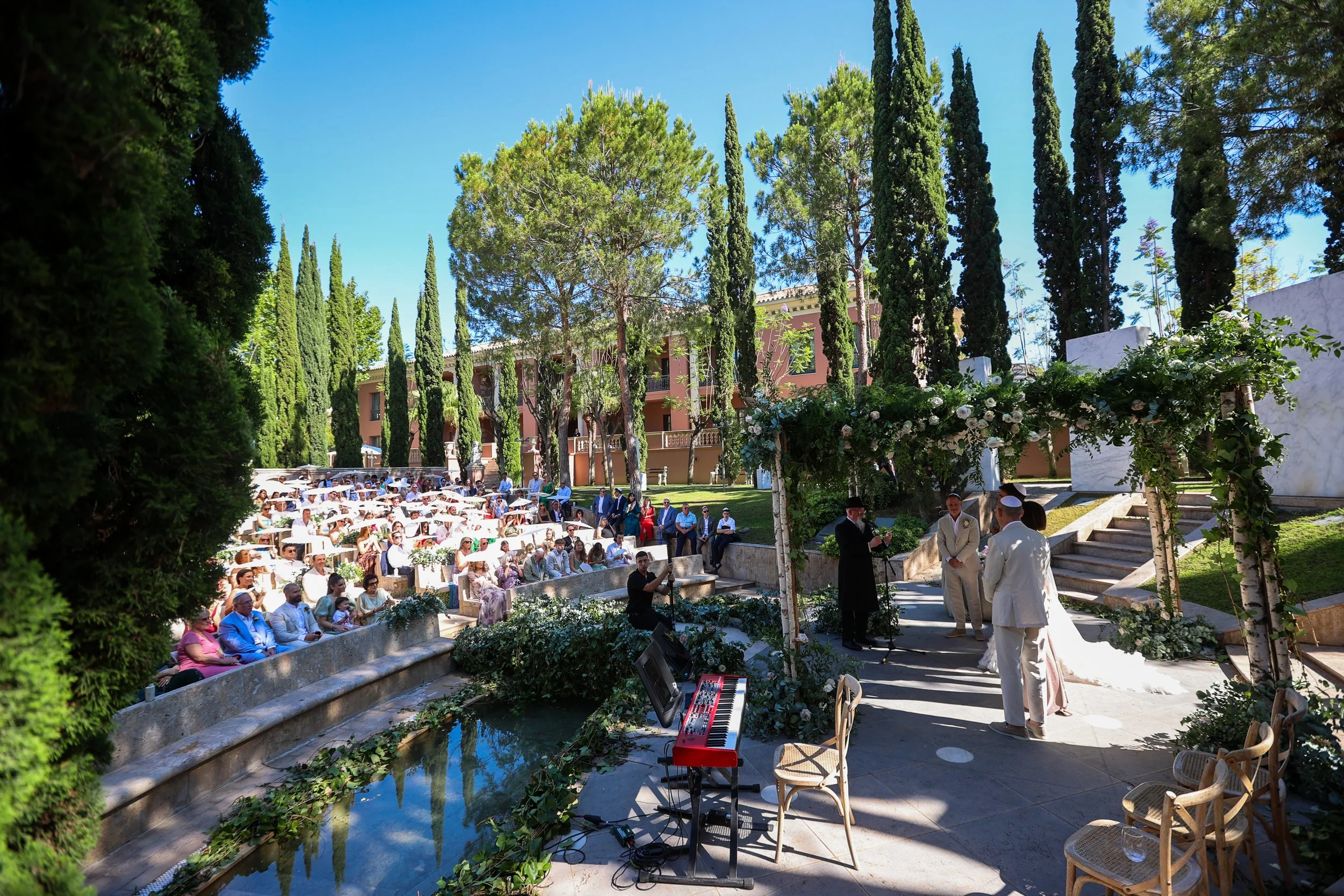 An outdoor wedding ceremony with guests seated on white chairs, surrounded by tall green trees and lush landscaping. The ceremony is taking place near a small water feature with flowers and greenery. Musicians are set up with a keyboard, and people a