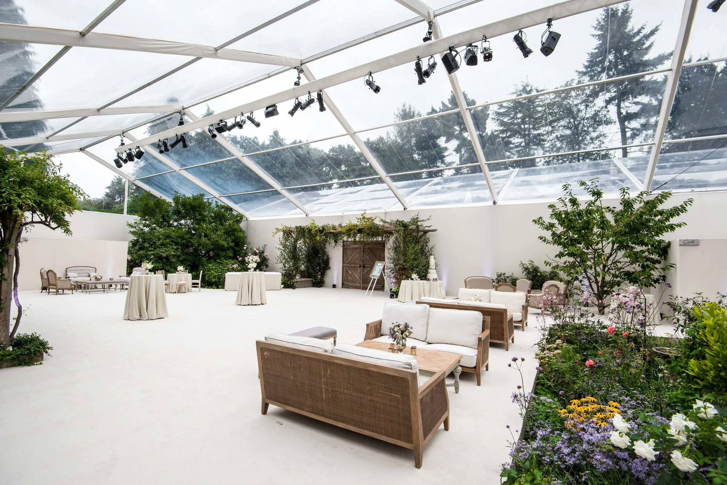 Indoor event space with white walls and ceiling, decorated with greenery and flowers, featuring seating areas with sofas and chairs, and tables with centerpieces, under a transparent roof with stage lighting fixtures.