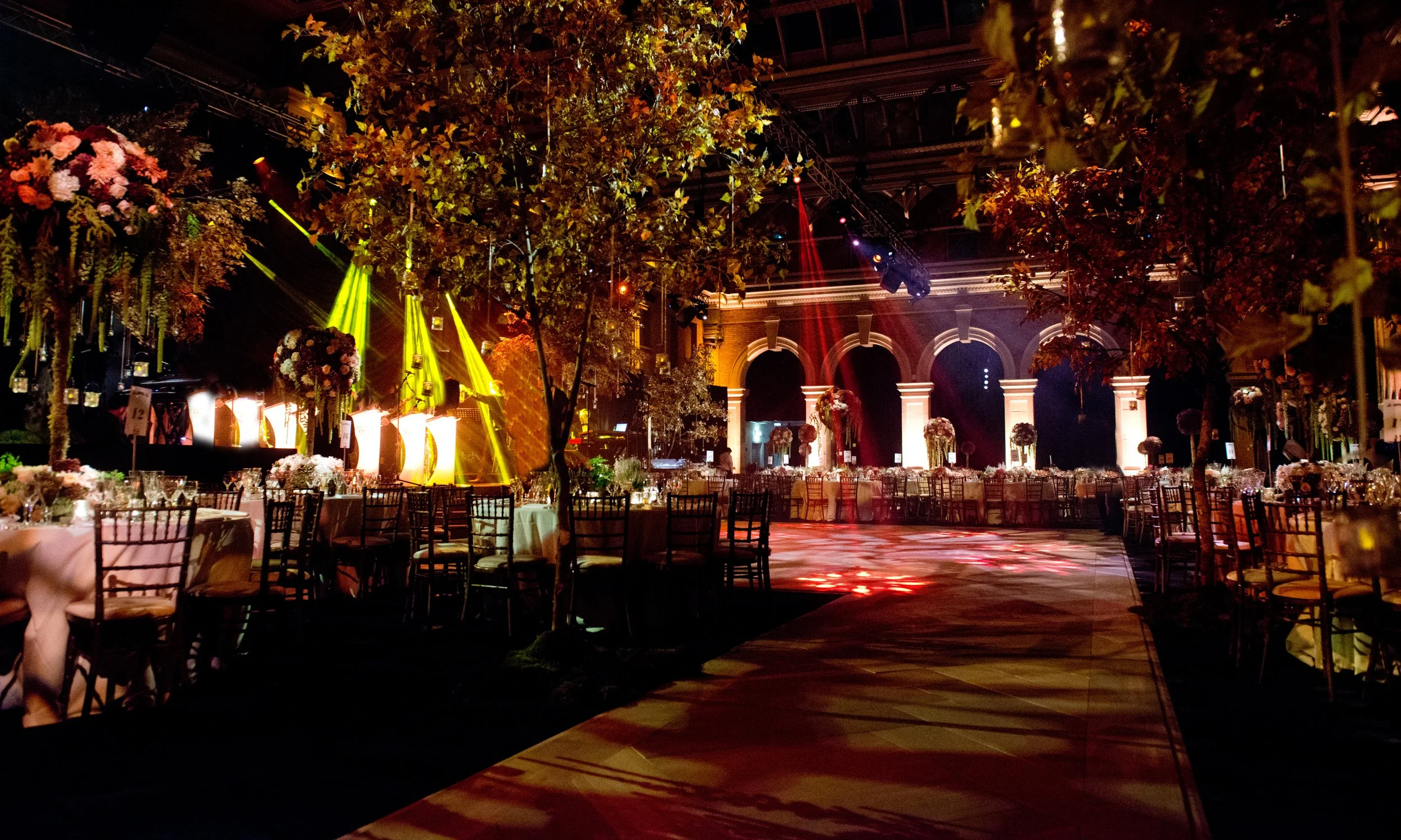 An elegantly decorated event hall with tables adorned with floral arrangements and white tablecloths, illuminated by colorful stage lights, featuring tall greenery and floral centerpieces, under a decorated ceiling with large arches in the background