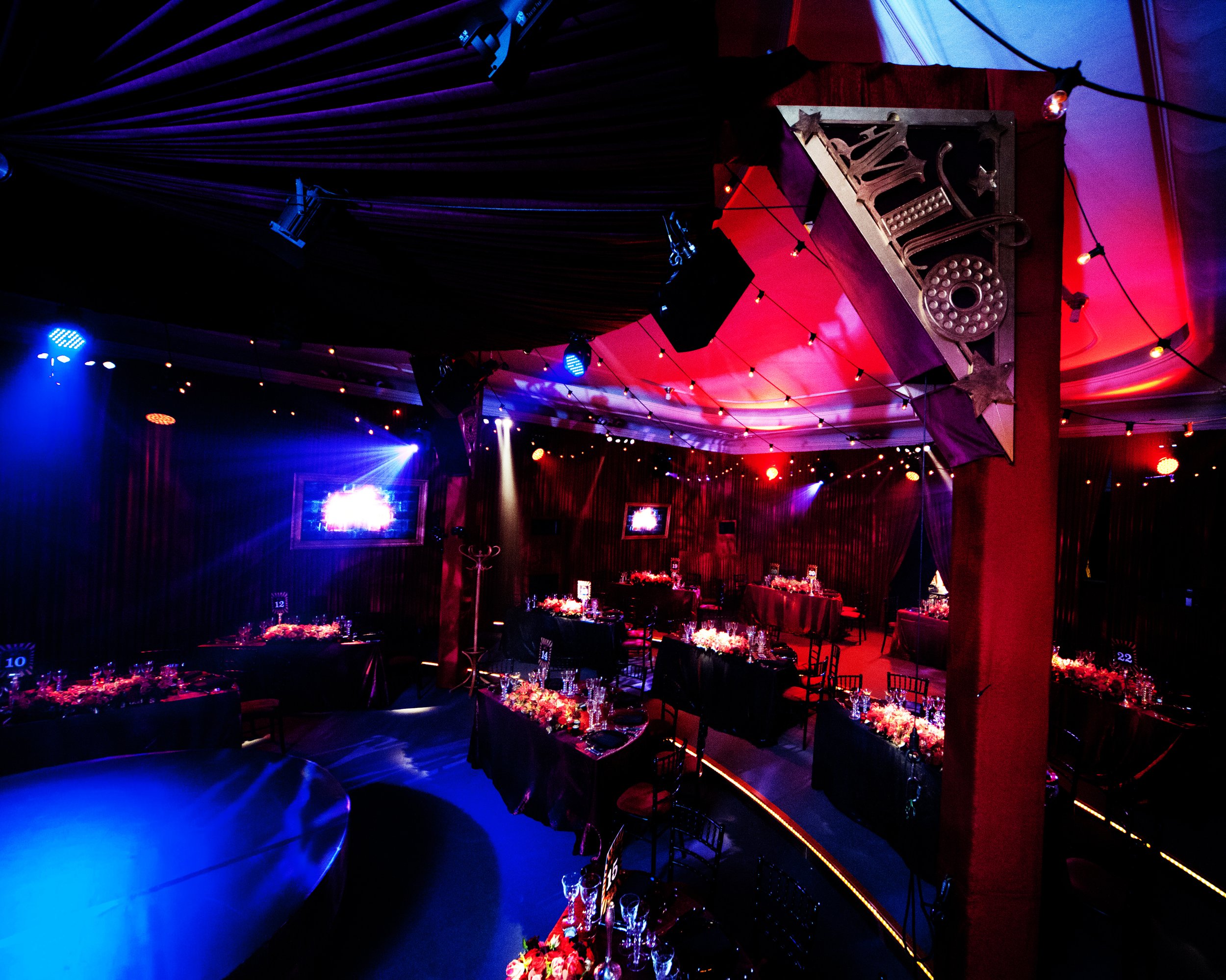 Decorated event space with dim lighting and colorful spotlights, featuring tables set for a celebration with flowers and glassware, and a stage with screens.