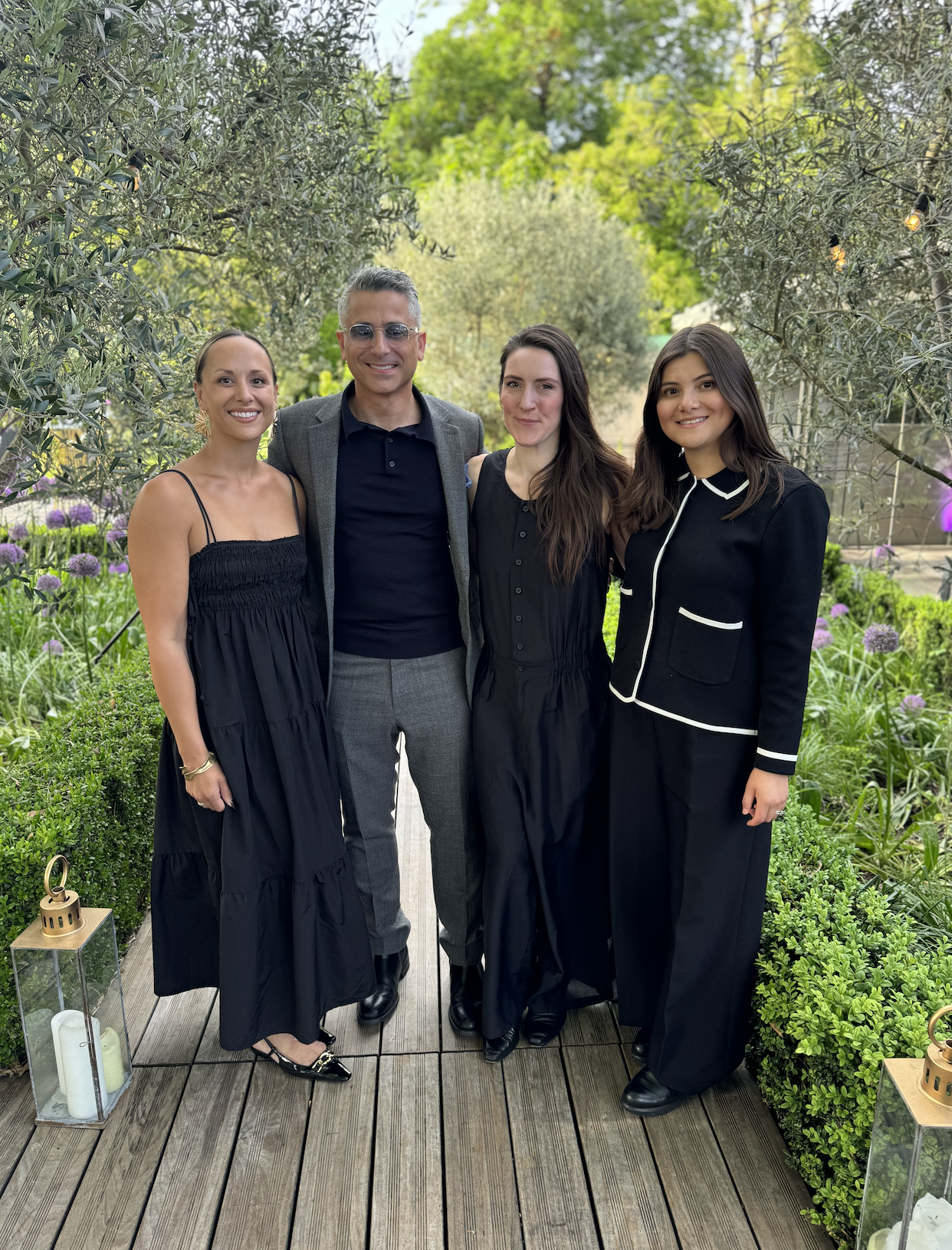 Four people standing on a wooden pathway in a garden, smiling at the camera. The group includes two women on the left and right, a man in the middle, and a woman on the right. The background has lush green trees and purple flowers.