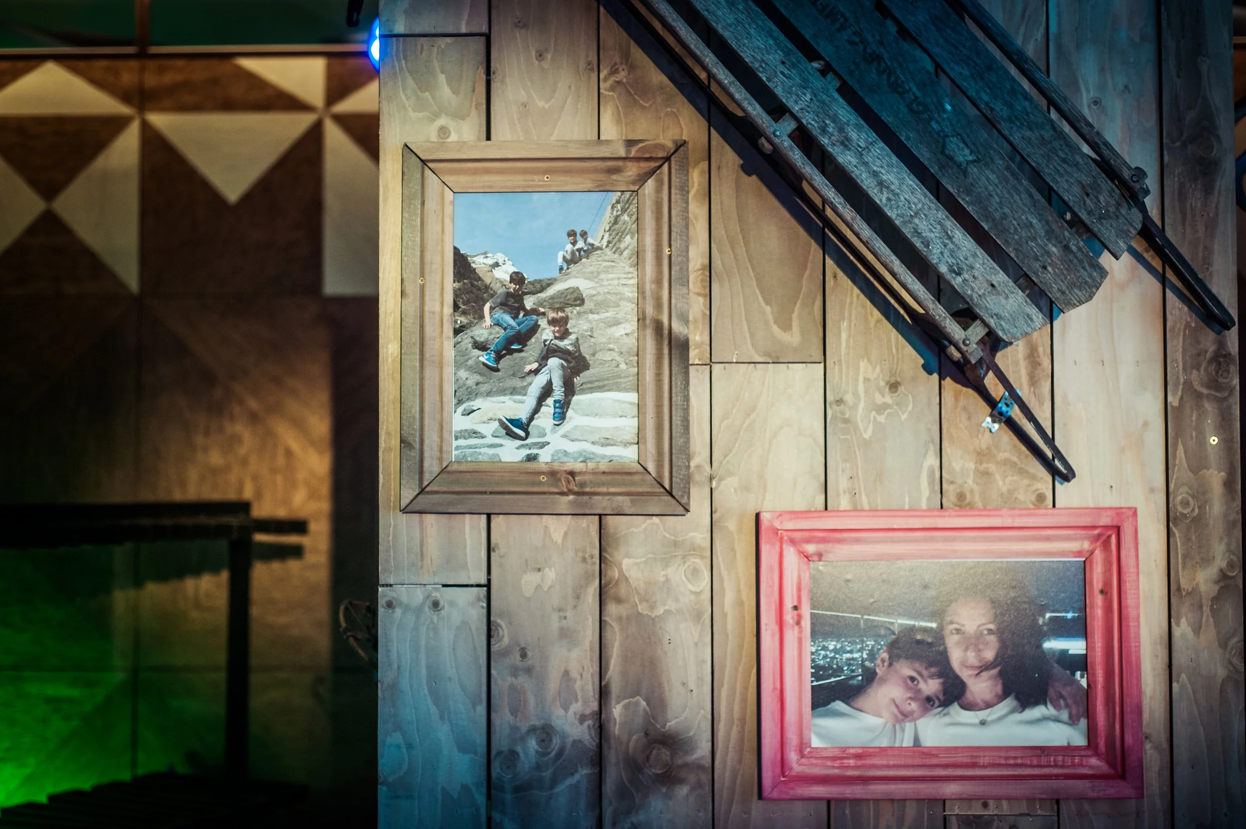 A framed photograph of two boys sitting on rocks outdoors, with a mountain landscape and blue sky in the background, hanging on a wooden wall with other photos and pictures nearby.