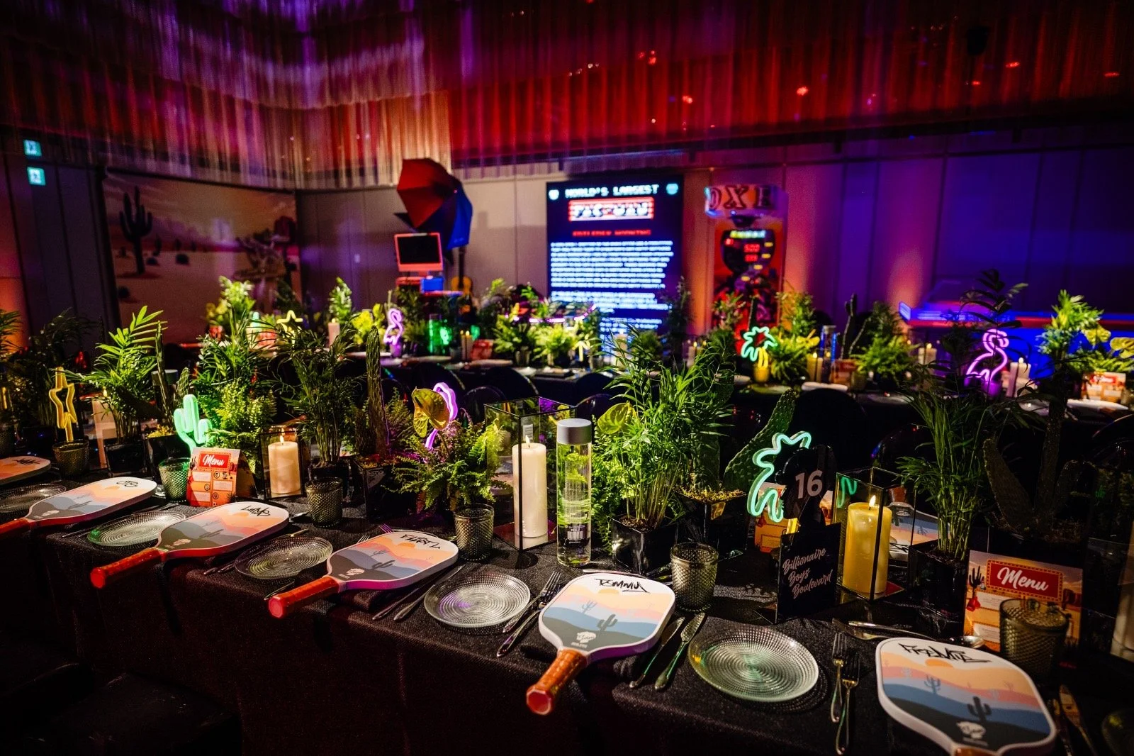 A decorated indoor dining area with tables set for a meal, featuring tropical plants, neon flamingo and palm tree signs, paddle shaped plates, and colorful lighting. There is a dance or game area in the background with a large digital screen and arca