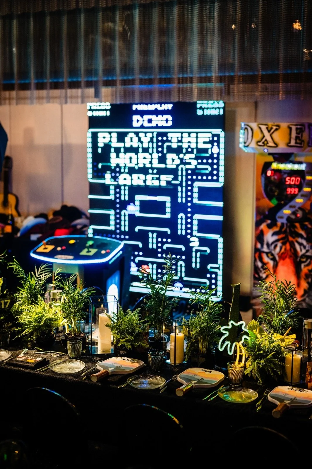 An arcade game screen displays Pac-Man and the words "Play the World’s Largest". The foreground features a table set with dining utensils, candles, and green plants. There is a neon palm tree sign and the edge of a food truck or booth with a tiger pi