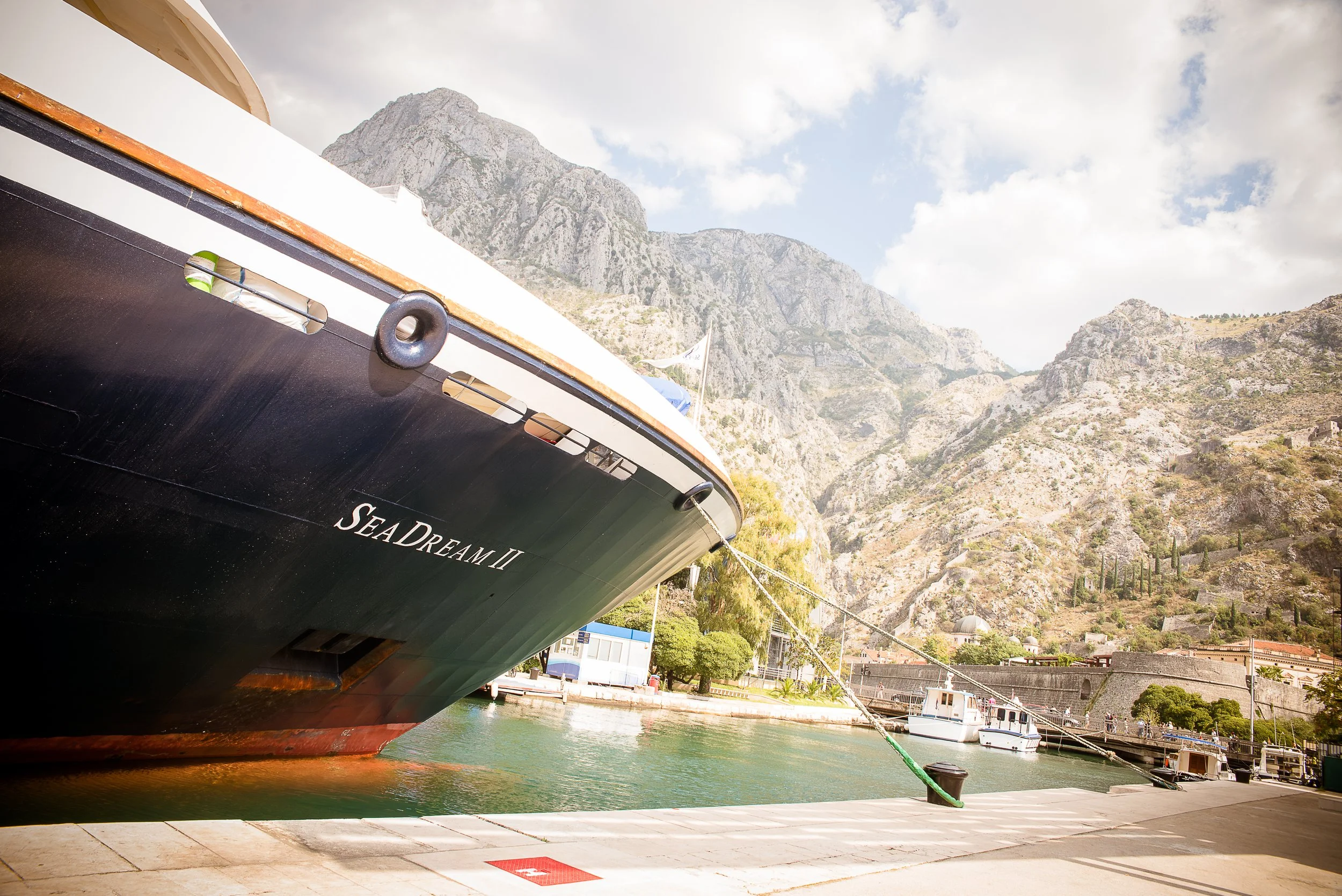 Close-up view of the bow of a boat named Sea Dream II, docked at a marina with water, mountains, and a partly cloudy sky in the background.