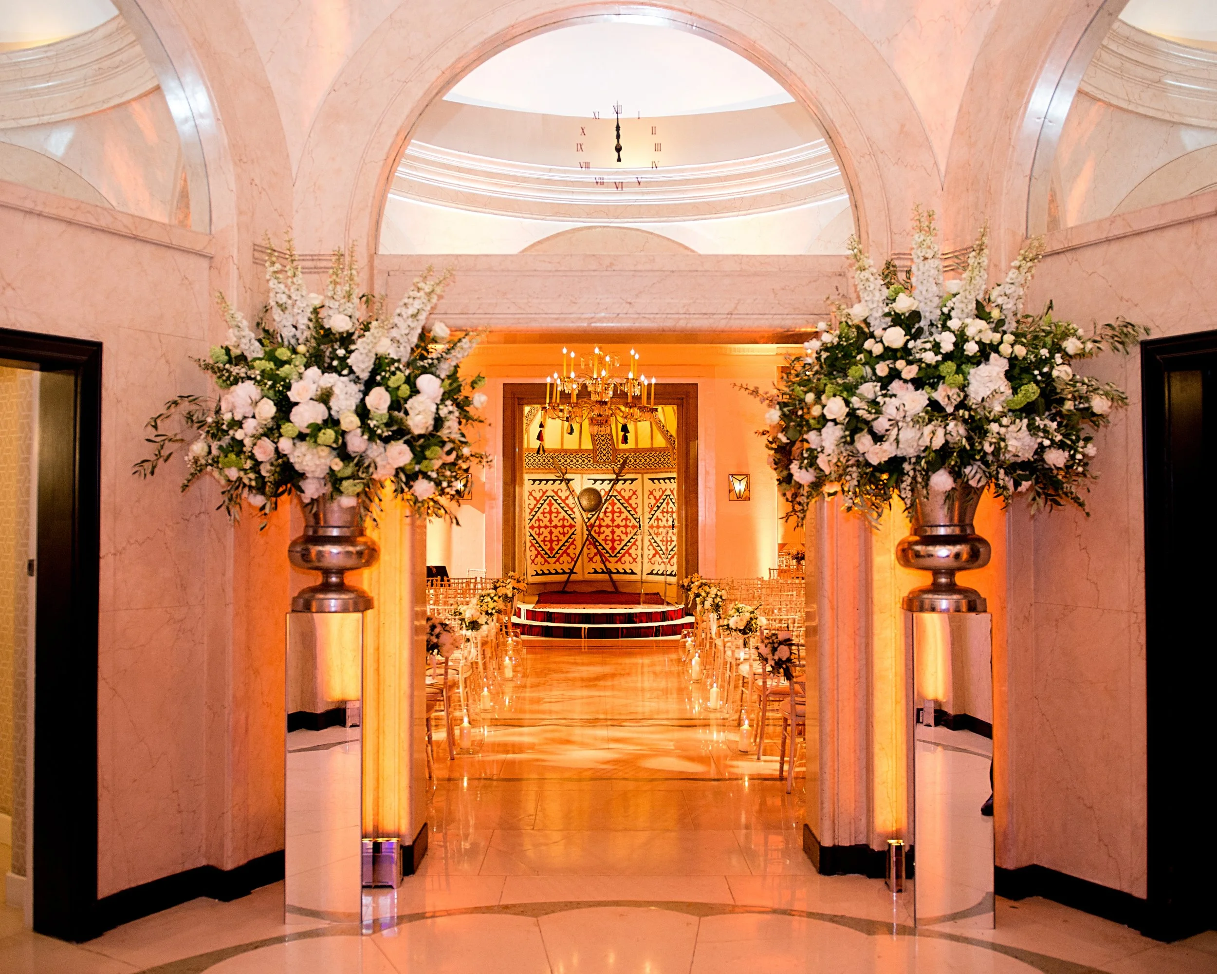 An elegant wedding venue with a marble hallway leading to an altar decorated with large floral arrangements and a chandelier, illuminated in warm lighting.