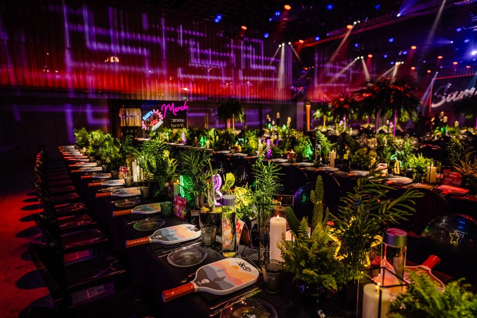 A festive indoor event decorated with a tropical theme, featuring lush green plants, candles, and neon signs, with long banquet tables set for dining under colorful lighting Bar Mitzvah