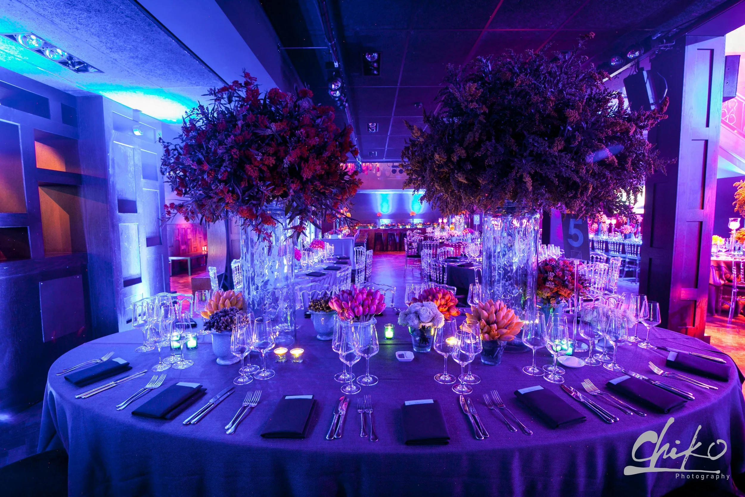 An elegantly decorated event space illuminated with purple and blue lighting, featuring round tables set with silverware, black napkins, various flower arrangements, and large floral centerpieces, with a stage in the background.