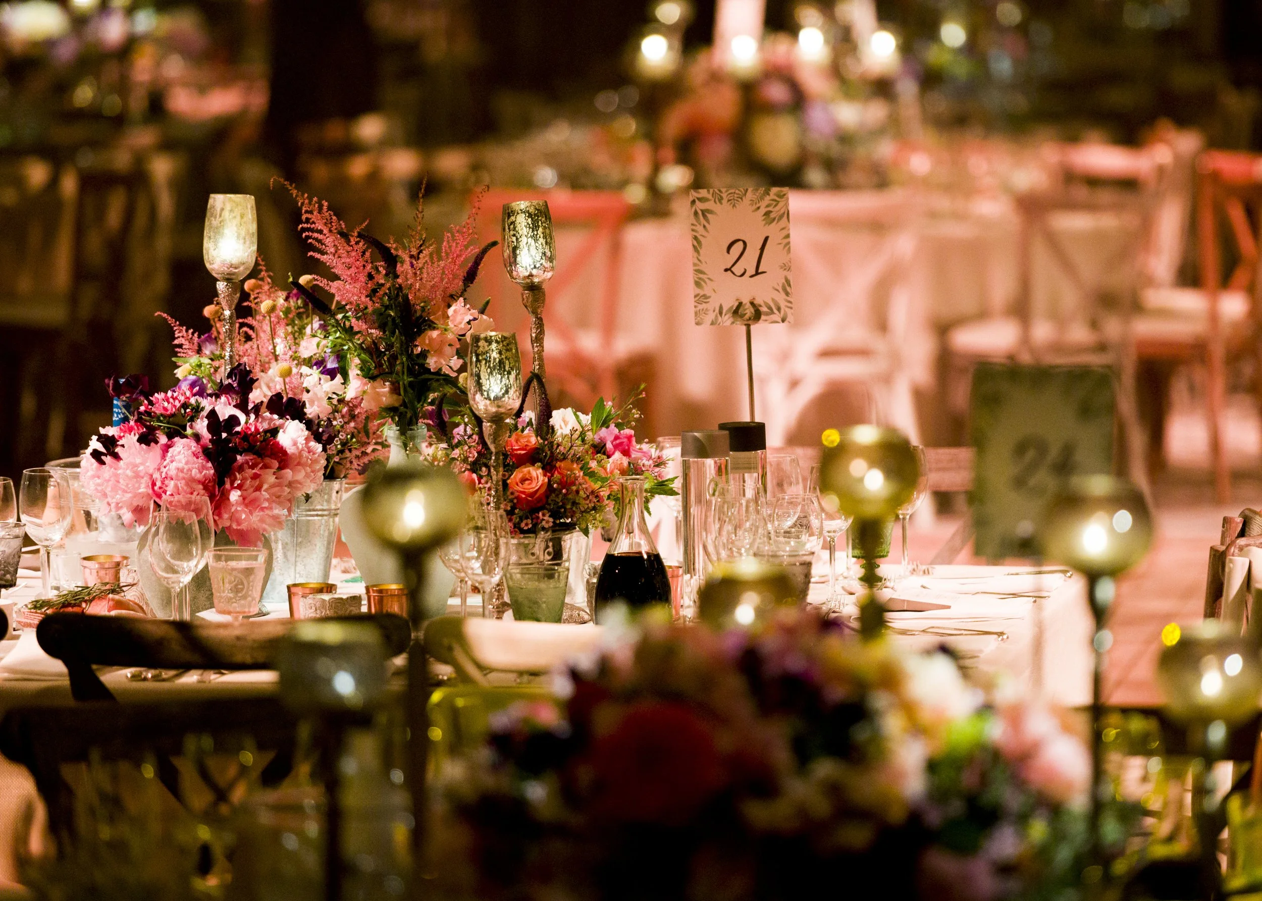 A decorated, floral centerpiece with pink and purple flowers, candles, and table number 21 on a wedding or event table, with blurred background and warm lighting.