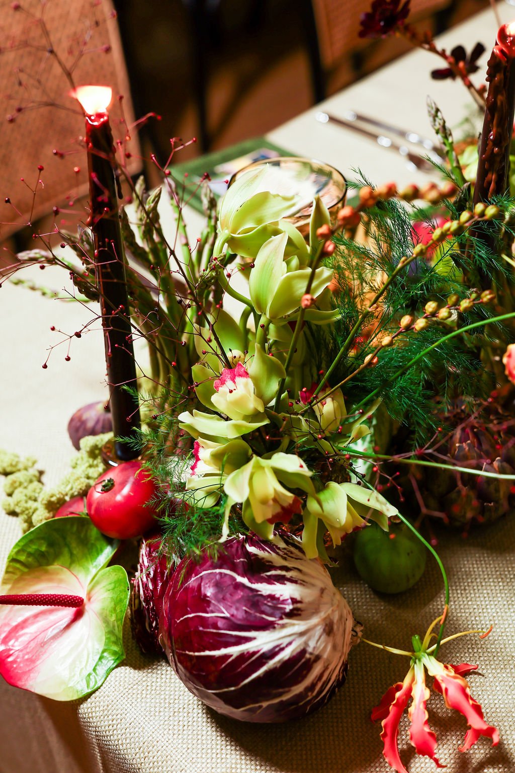 A floral centerpiece with white orchids, an anthurium, and other colorful flowers and greenery, arranged in vases on a dining table with cutlery and napkins.