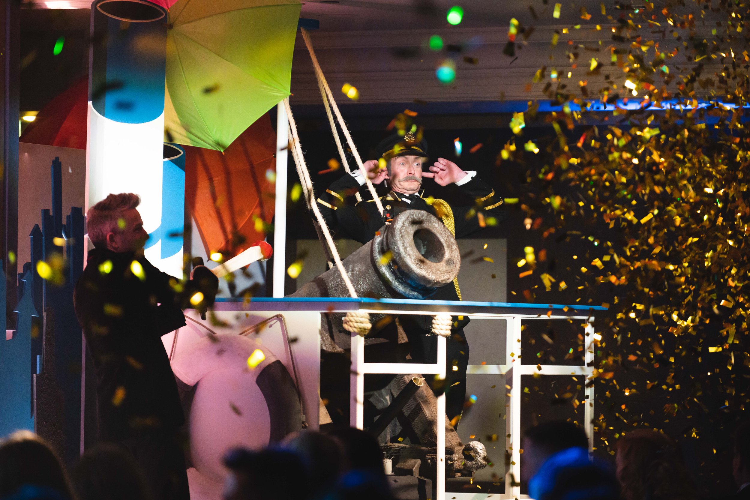 A man dressed as a captain on a boat-themed float during a parade, surrounded by falling gold confetti and colorful umbrellas, with a crowd watching.