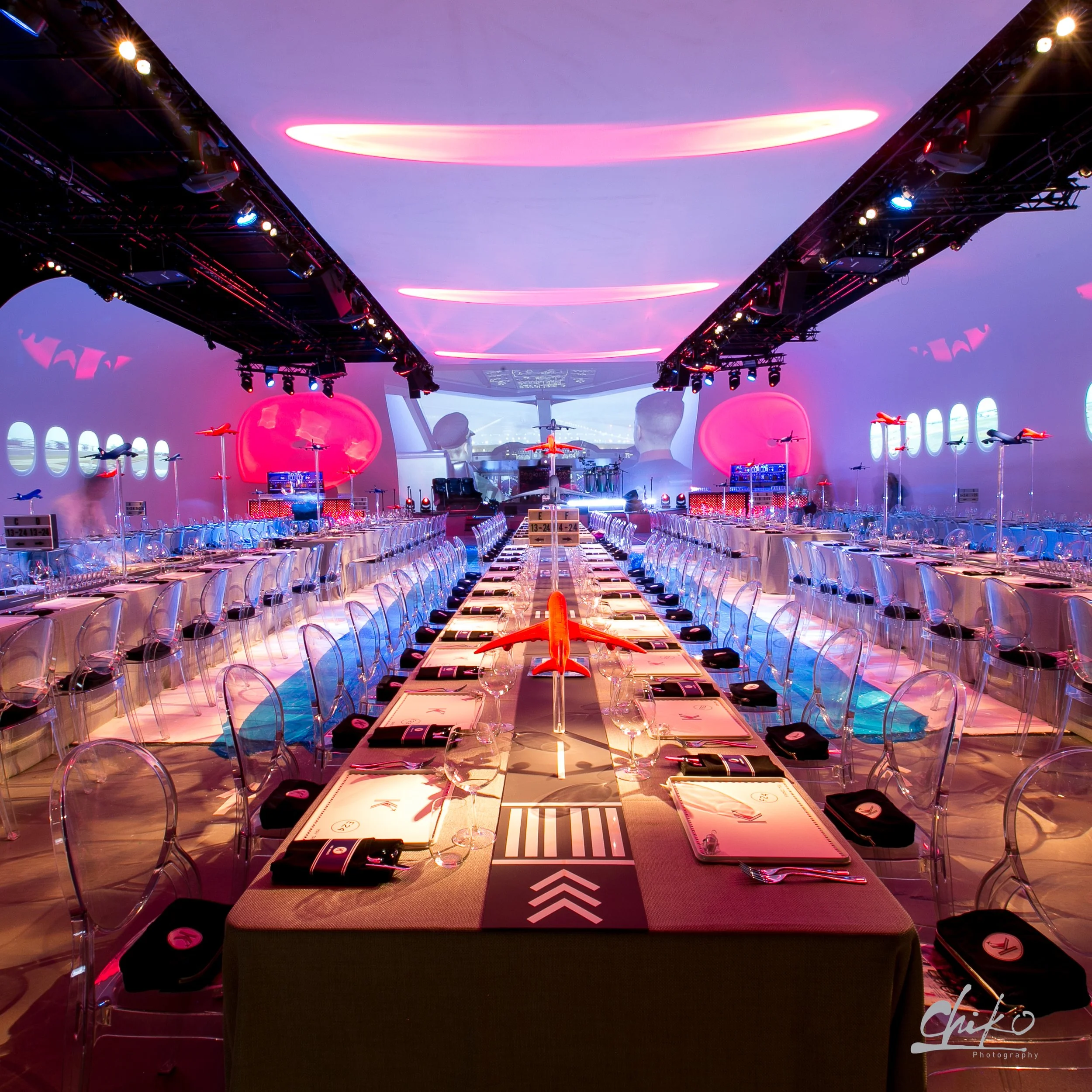 A banquet or conference hall with a long table set for dining, illuminated with colorful neon and stage lighting, featuring chairs, water glasses, and place settings, with a large screen and lighting equipment above.