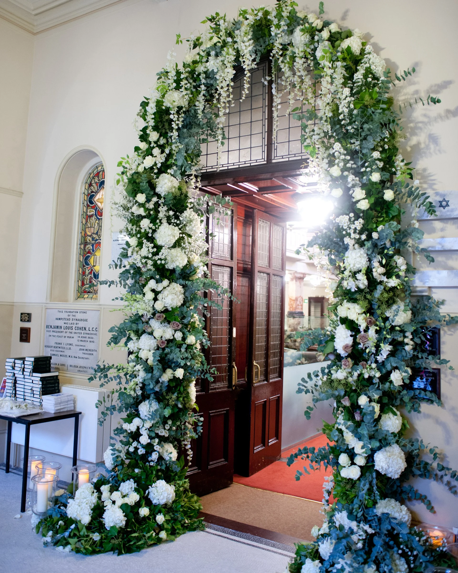 Floral archway decorated with white and light purple flowers, greenery, and candles in glass holders, set up inside a building near a wooden door.