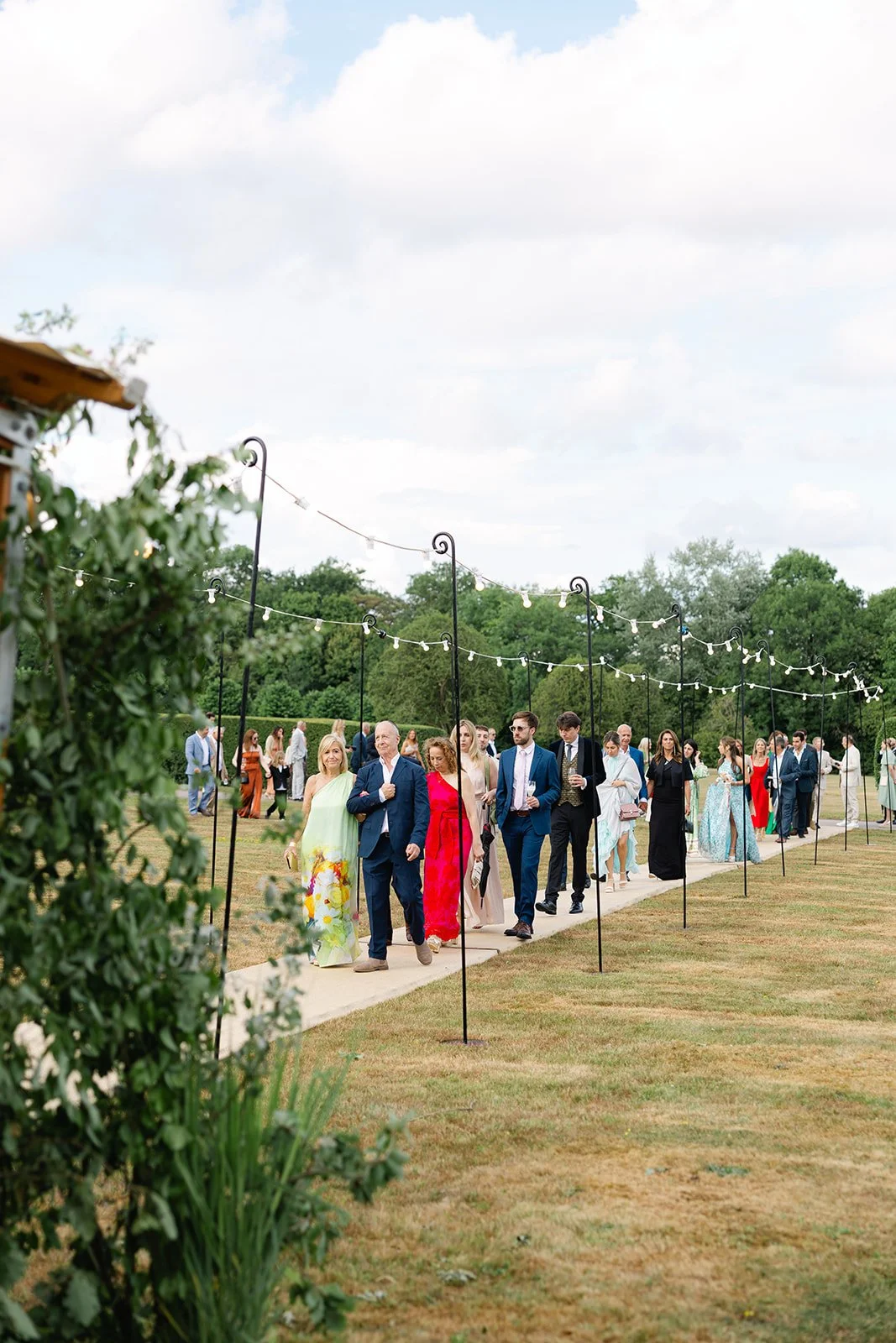 People in formal attire walking along an outdoor pathway decorated with string lights at a garden wedding reception.