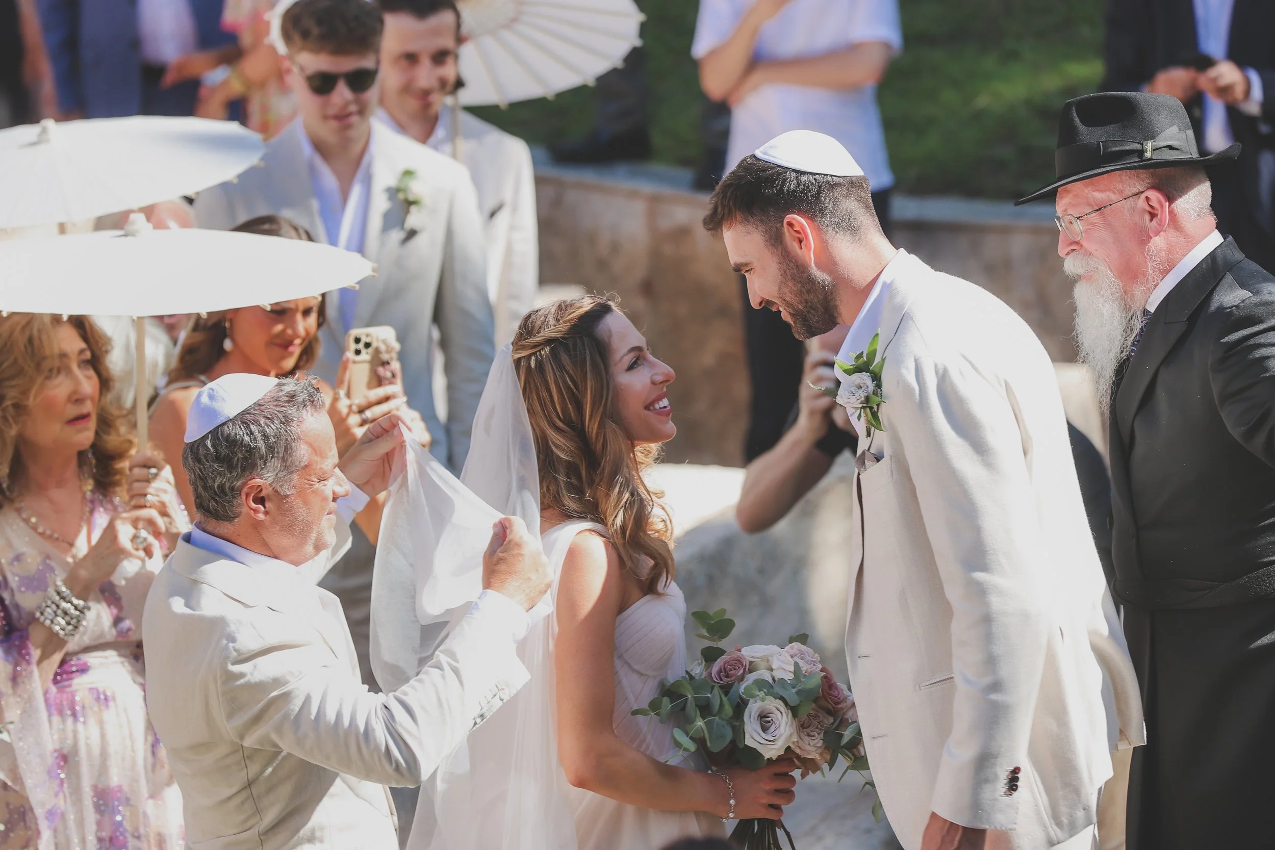 A wedding ceremony with a bride and groom exchanging vows, surrounded by family and friends. The bride is holding a bouquet of flowers, and the groom, wearing a white jacket and a yarmulke, is leaning towards her. Guests are dressed in formal attire,