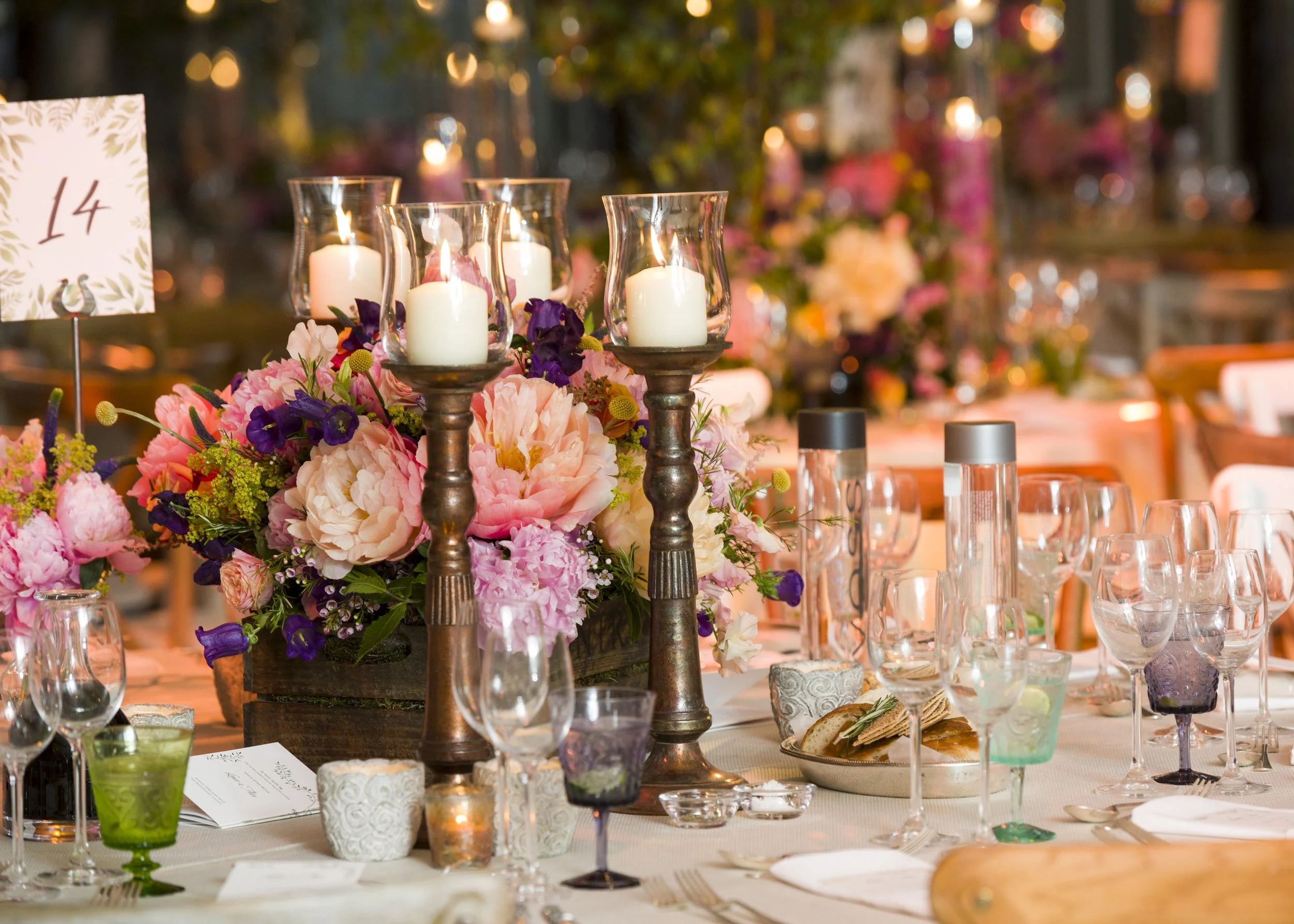 A decorated event table with flowers, candles in glass holders, glassware, and table settings, with a blurred background of similar decorations and warm lighting.