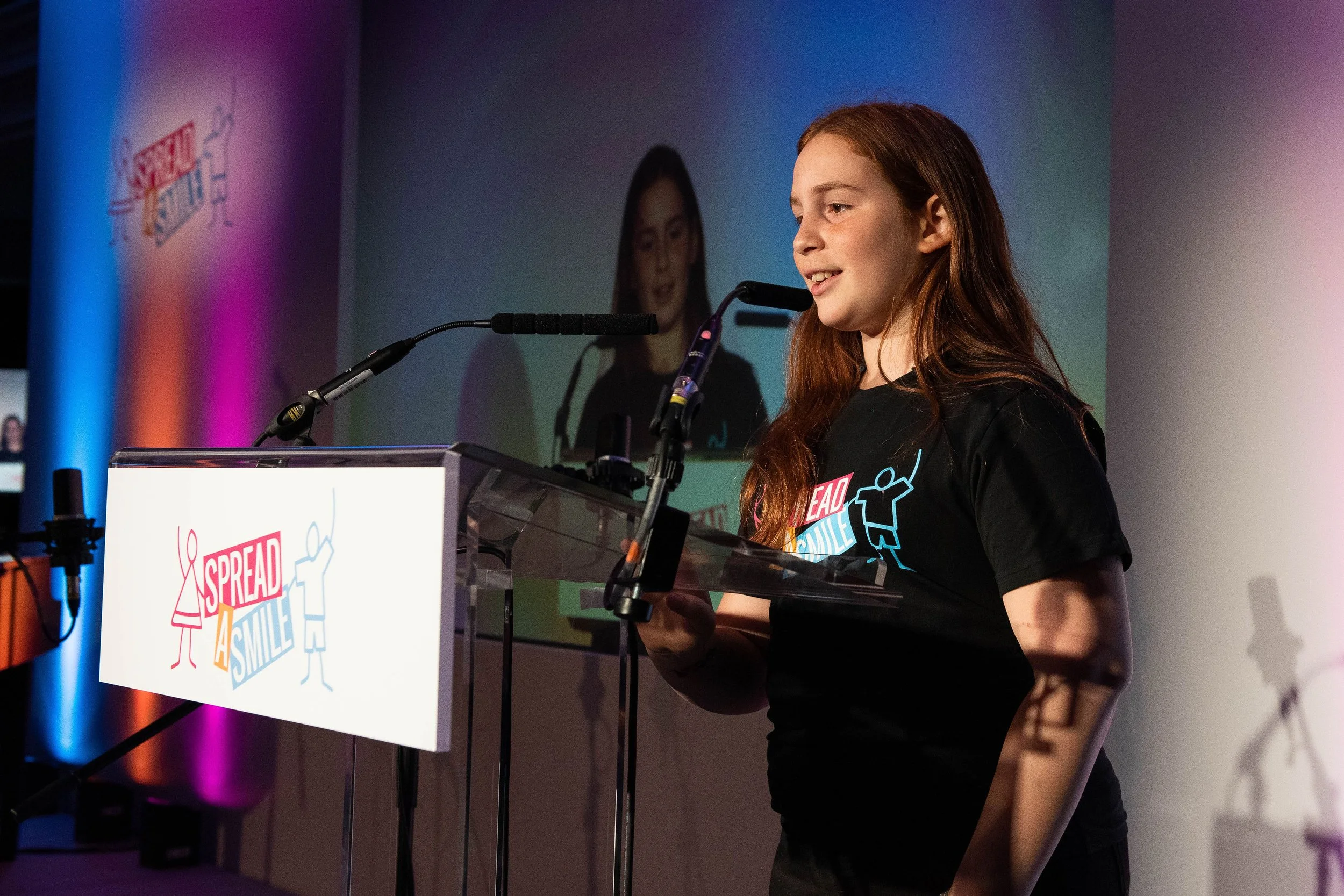A young woman with red hair is speaking at a podium during an event. She is smiling and looking to her right. She is wearing a black T-shirt with a graphic design. There are two microphones on the podium, and a large screen behind her displays her face. The podium has a colorful logo that says Spread the Smile.