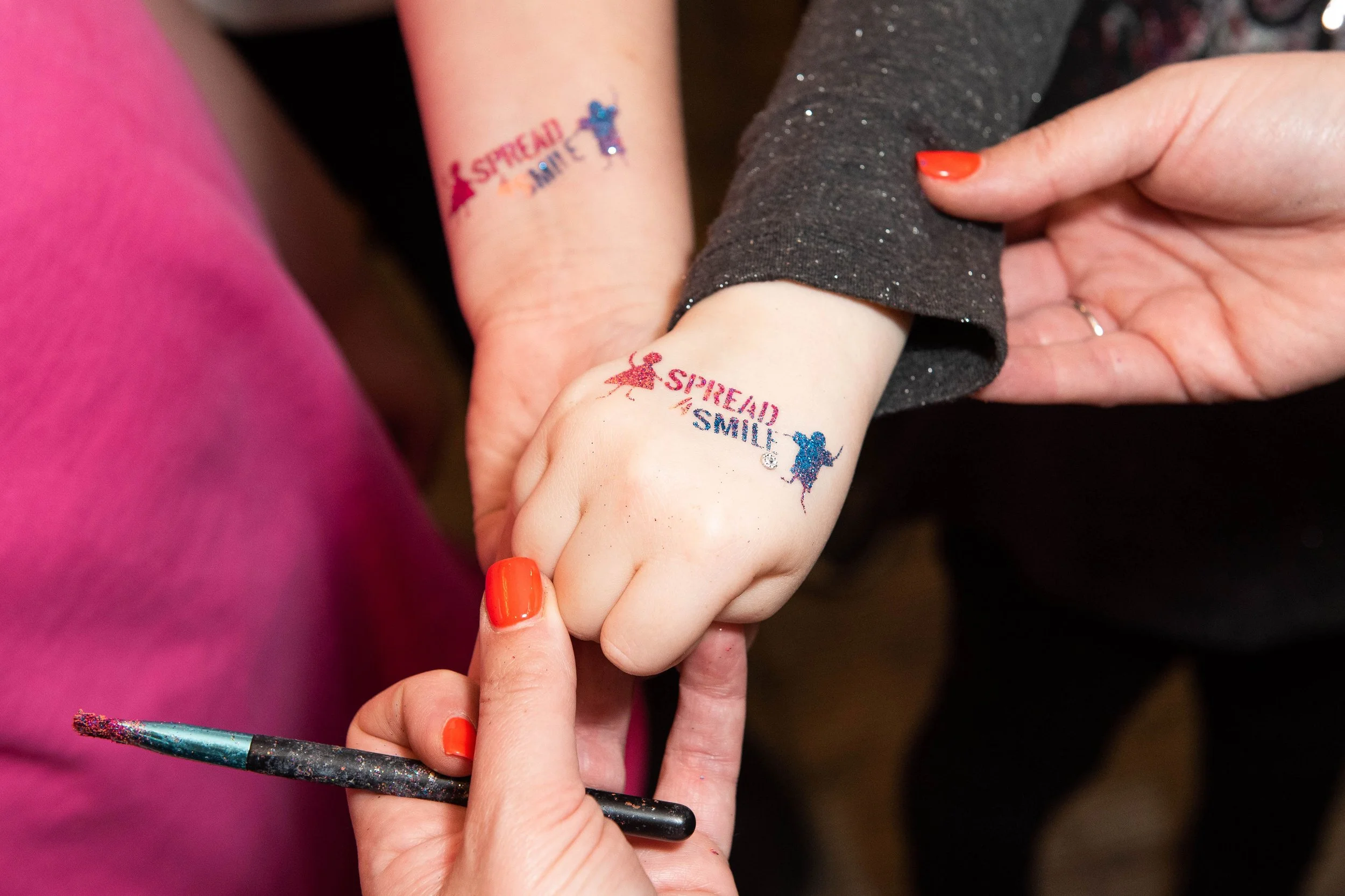 Close-up of two hands with red nail polish holding hands, with temporary tattoos on the wrists that say "STRENGTH AND SMILE" and feature a blue mouse and a red girl. A makeup brush with colorful paint is also visible.