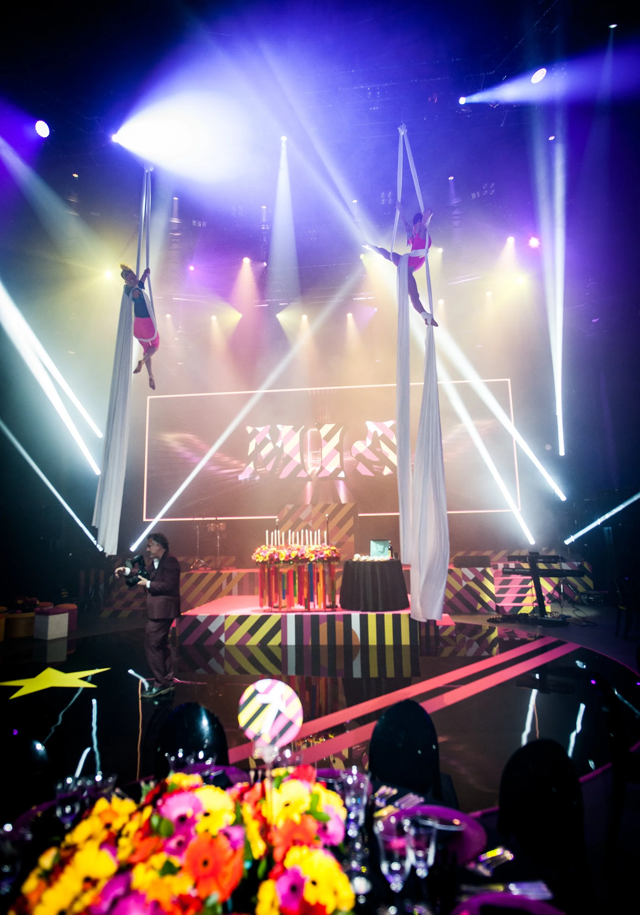 A brightly lit stage with aerial dancers on hanging silks, colorful stage designs, and a table with a birthday cake, floral arrangements, and balloons.  Bat Mizvah the roundhouse