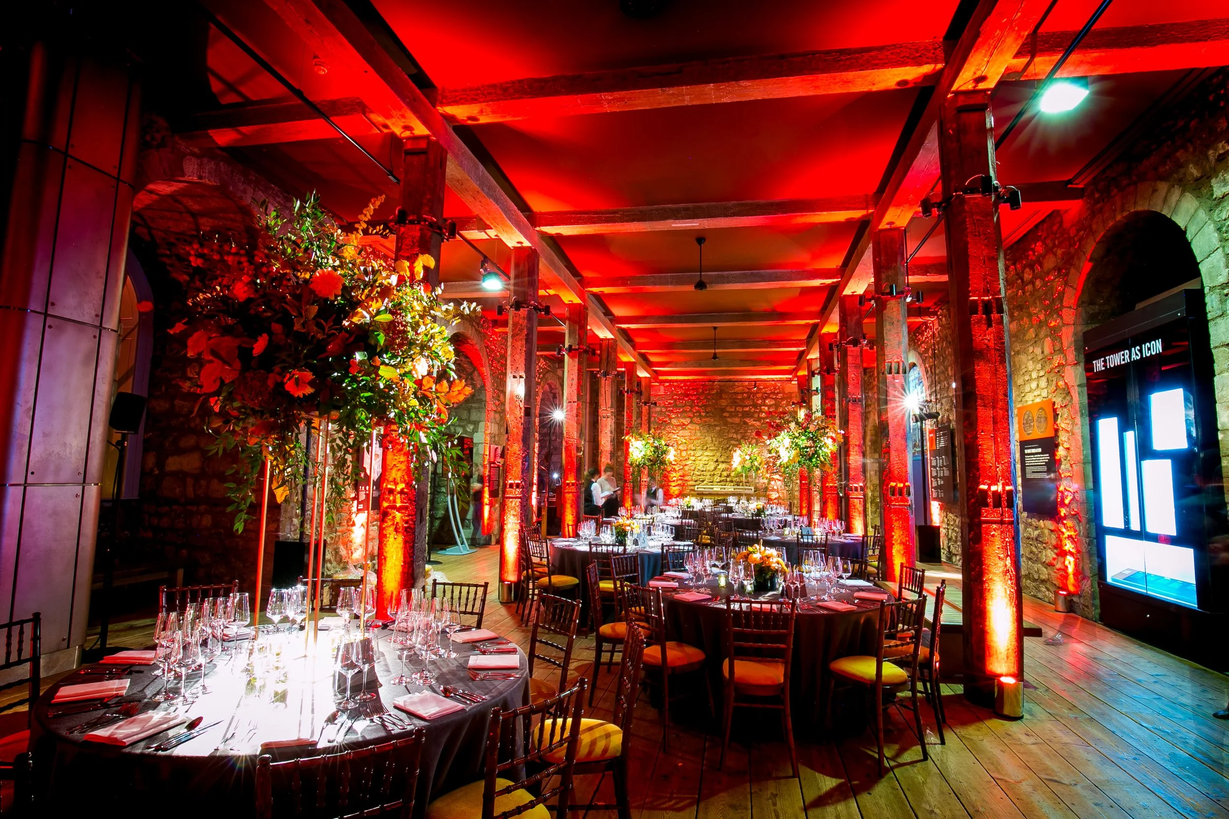 An indoor event space with exposed brick walls and wooden beams, illuminated with red lighting, decorated with large floral arrangements, and set up with round tables covered in black tablecloths and glassware.