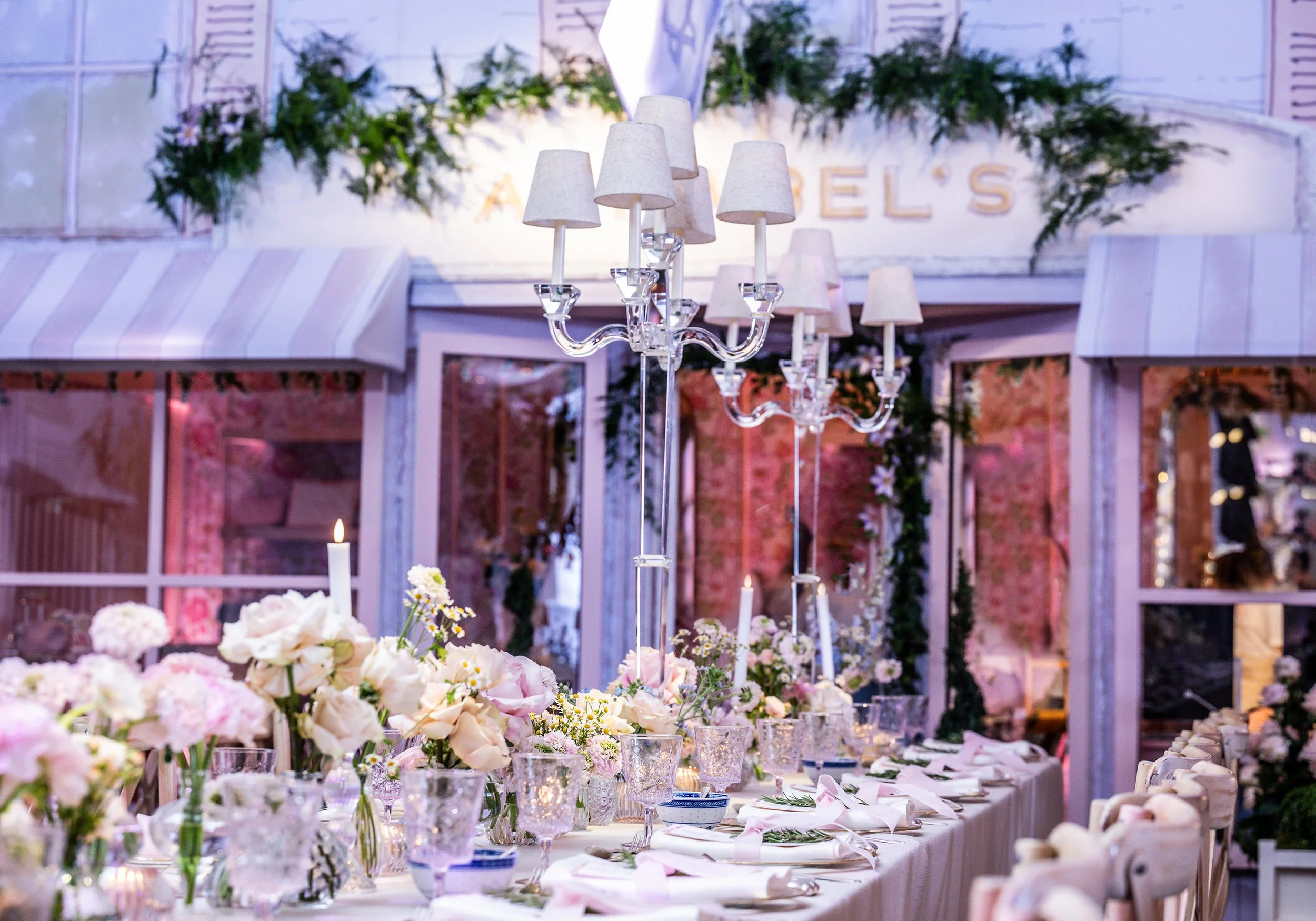 Elegant banquet table decorated with pink and white flowers, glassware, and candles. A crystal chandelier with white lampshades hangs above. Green greenery and flowers are in the background, with a sign that reads 'Angel's' partially visible.