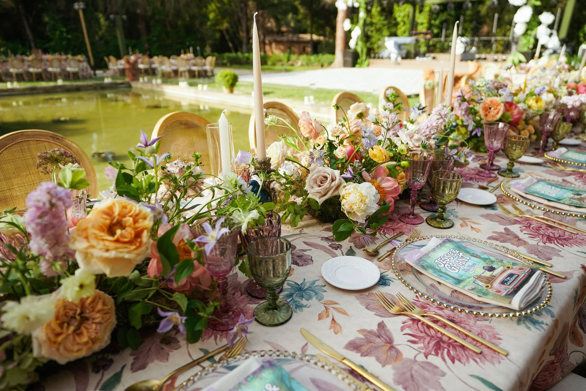 A long outdoor dining table decorated with a colorful floral tablecloth, arranged with pastel-colored flowers in various glasses, candles, and place settings, set in a lush garden environment.