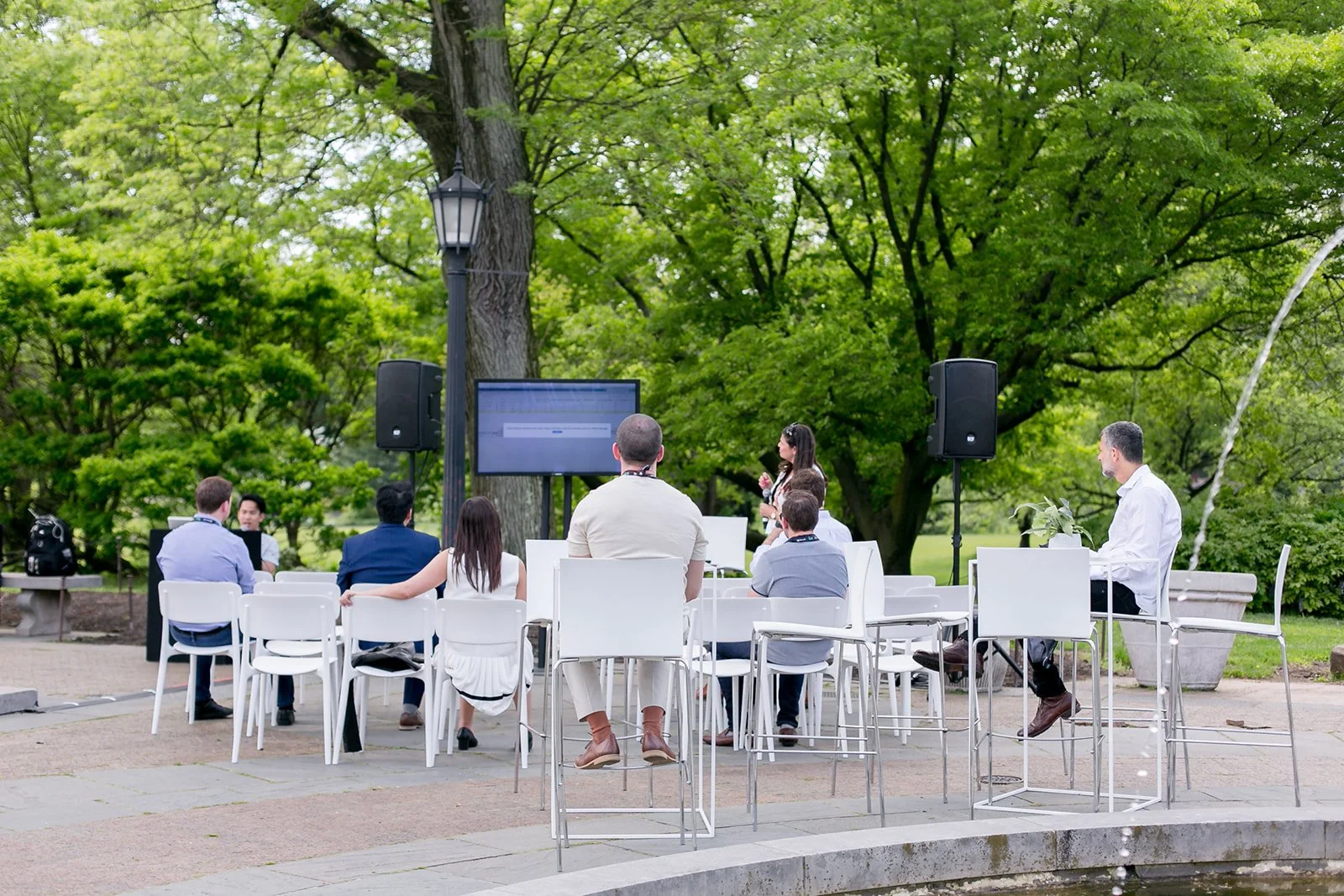 A outdoor meeting or presentation taking place in a park with people sitting on white chairs facing a large screen. A woman stands near the screen speaking to the group, with a laptop nearby. The setting is lush with green trees and a lamp post.