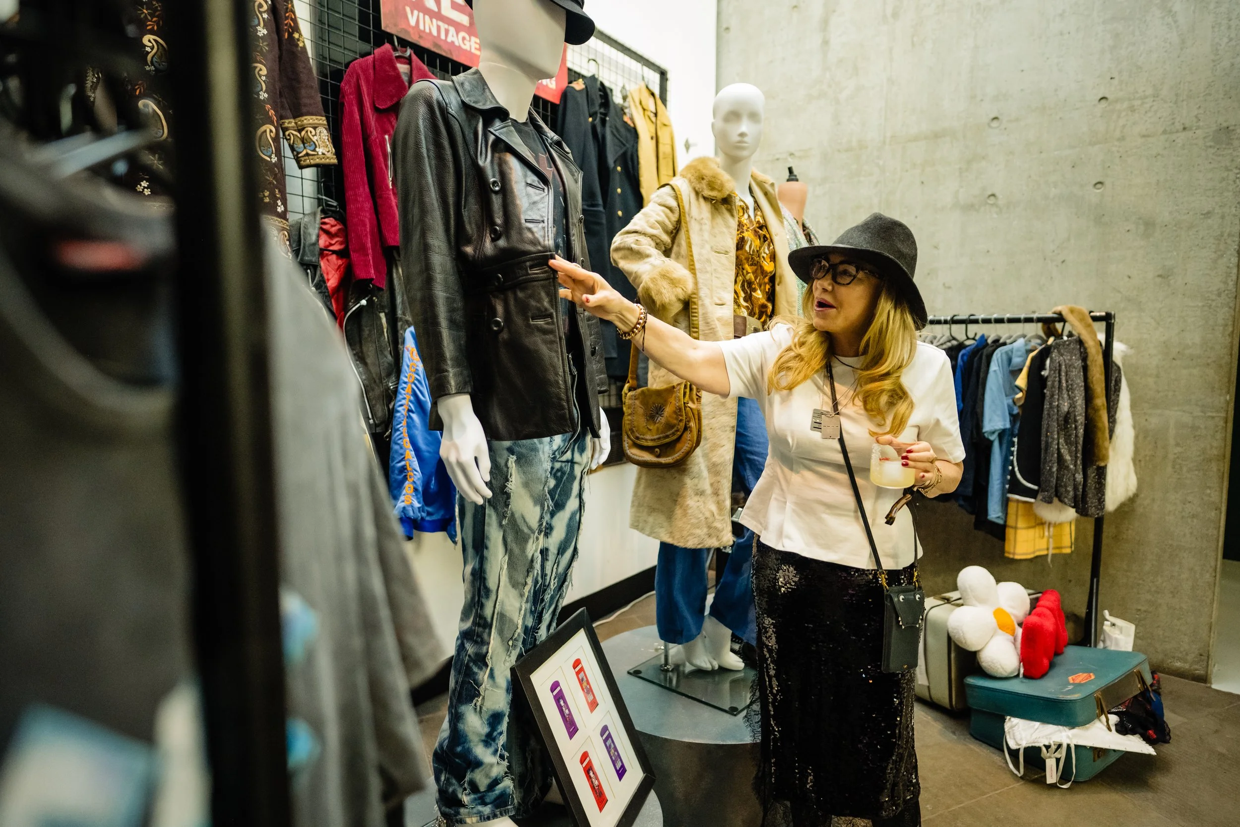 A woman with glasses and a hat shopping at a vintage clothing store, looking at a black leather jacket on a mannequin. There are racks of clothing and signs in the background.