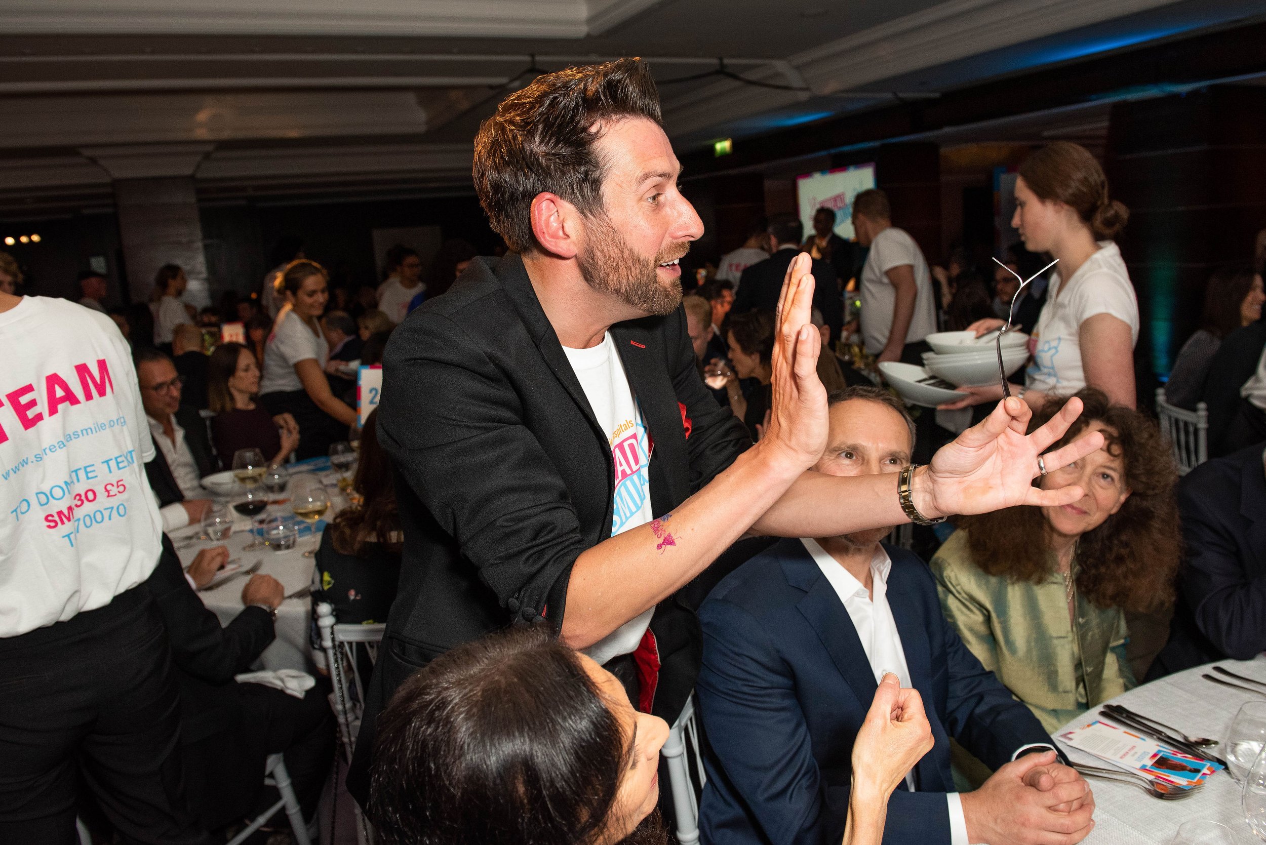 A man with a beard and styled dark hair raising his hand, engaging with an audience at a formal event. He is wearing a black blazer and white T-shirt. Several other people are seated at tables, and there are waitstaff serving food in the background.