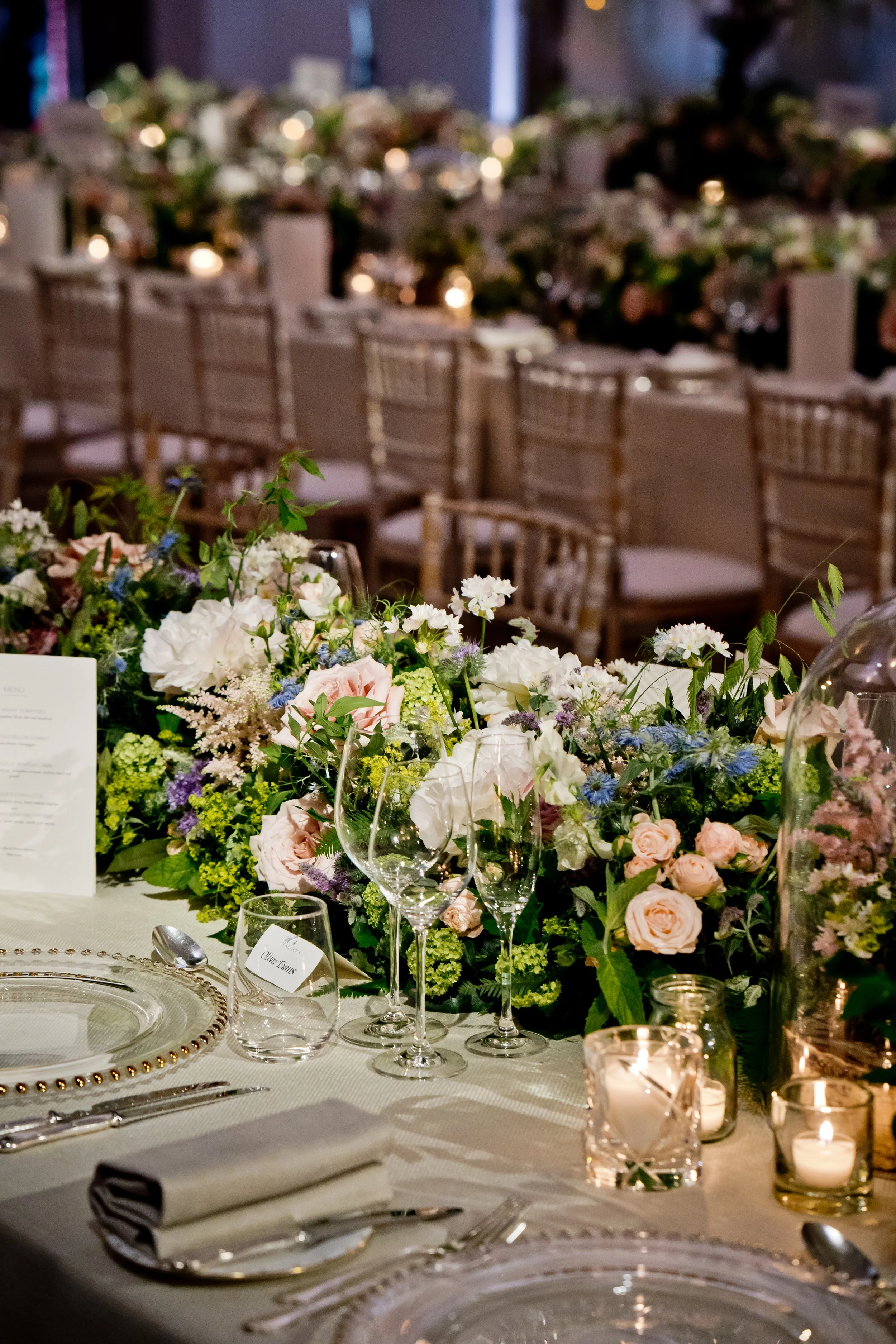 Elegant dining table with floral centerpiece, glassware, candles, and place settings in a decorated event space with chairs and subdued lighting.