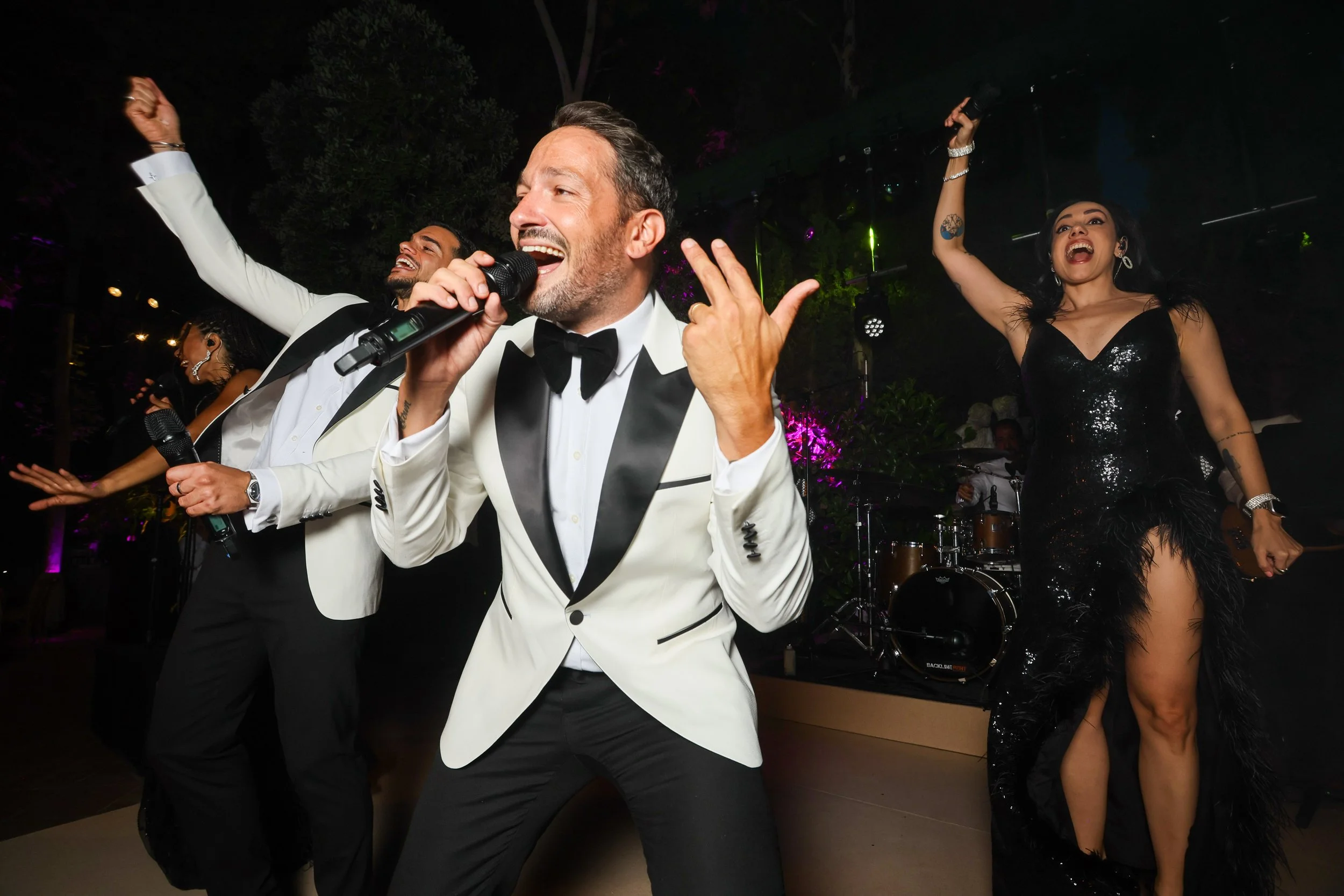 Happy man in a tuxedo singing into a microphone while dancing at a celebration, with women in glamorous dresses dancing in the background at night.