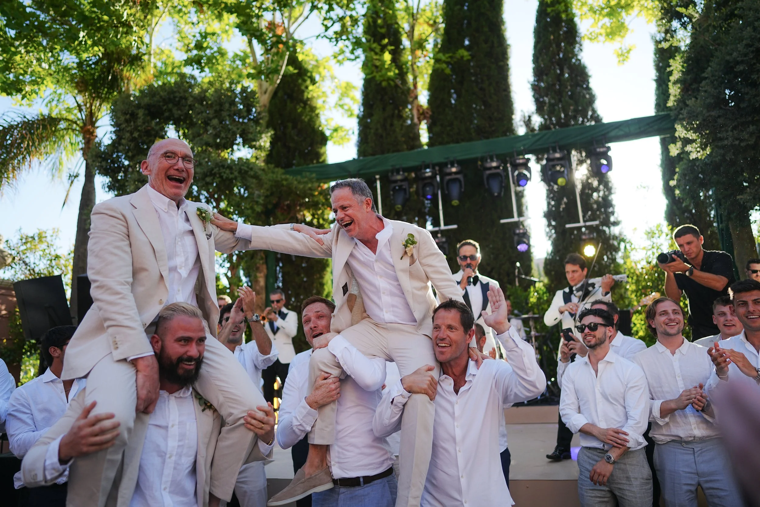 Group of men in white suits celebrating outdoors, with some sitting and lifting others on their shoulders, under tall trees with a green canopy, during daytime.